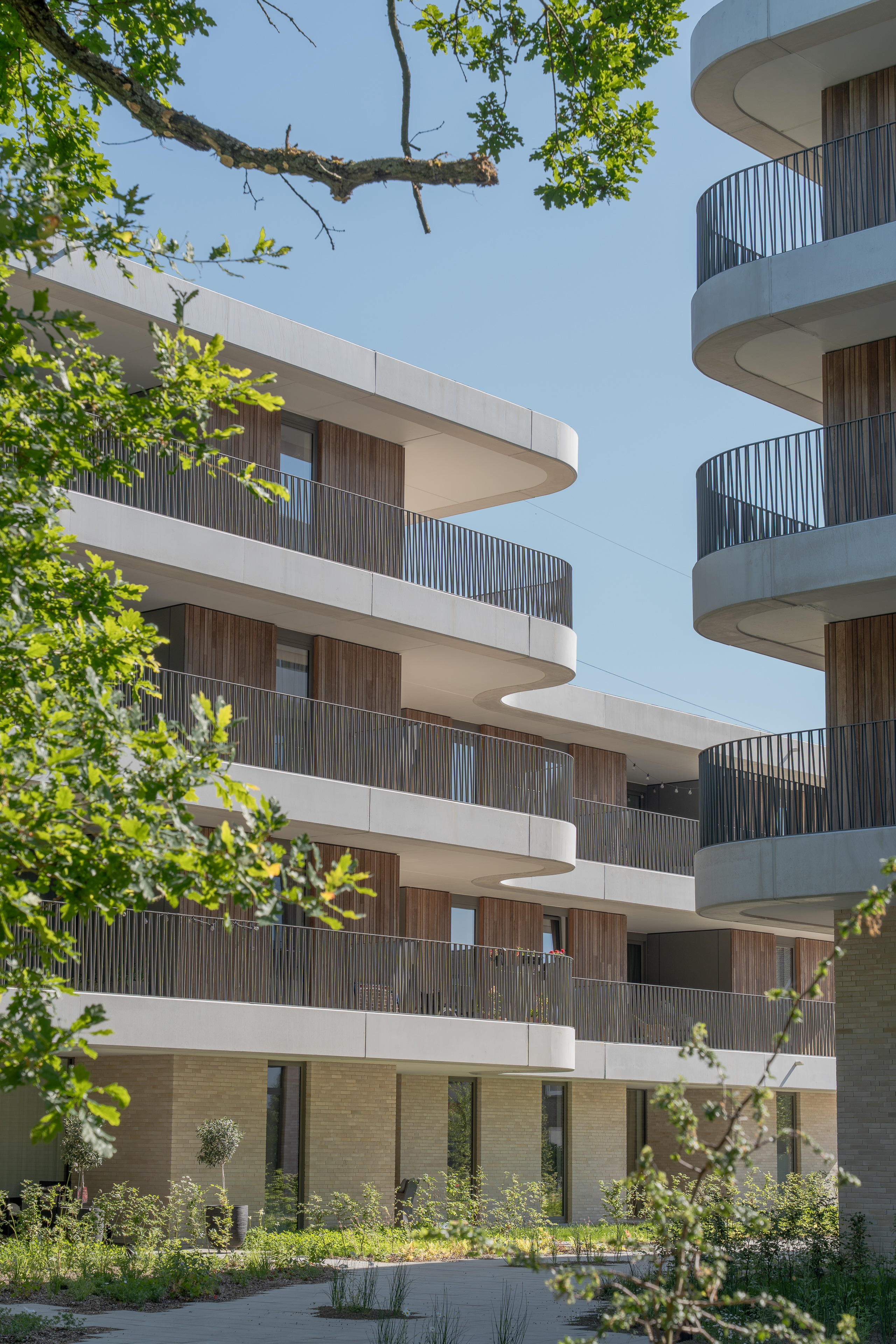 Twee moderne gebouwen met golvende balkons, gezien tussen bomen.