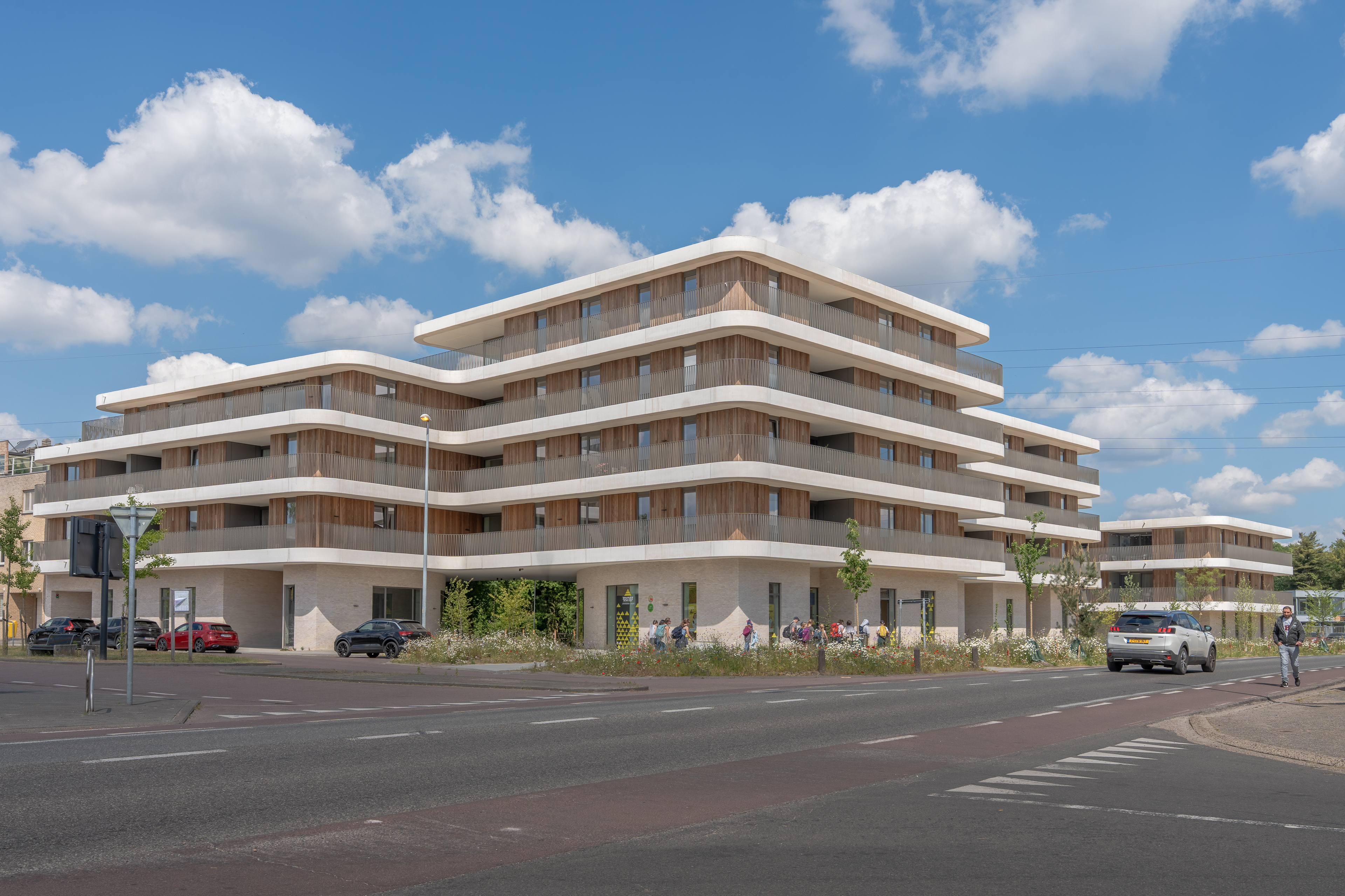 Straatzicht van groot appartementencomplex met afgeronde balkons, auto’s en voetgangers.