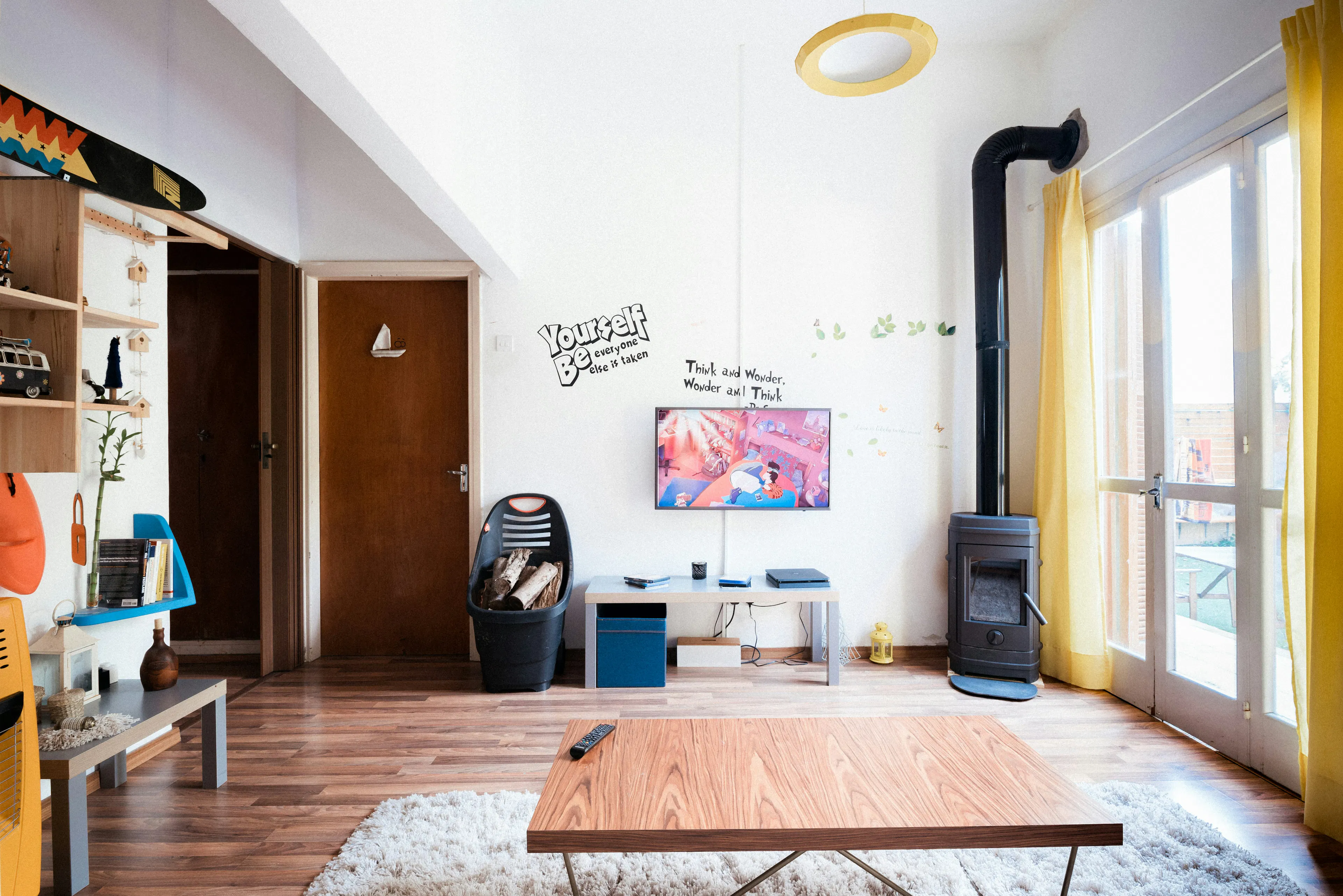 Bright modern living room with wood flooring, a coffee table, wall-mounted TV, and a black wood-burning stove near yellow curtains.