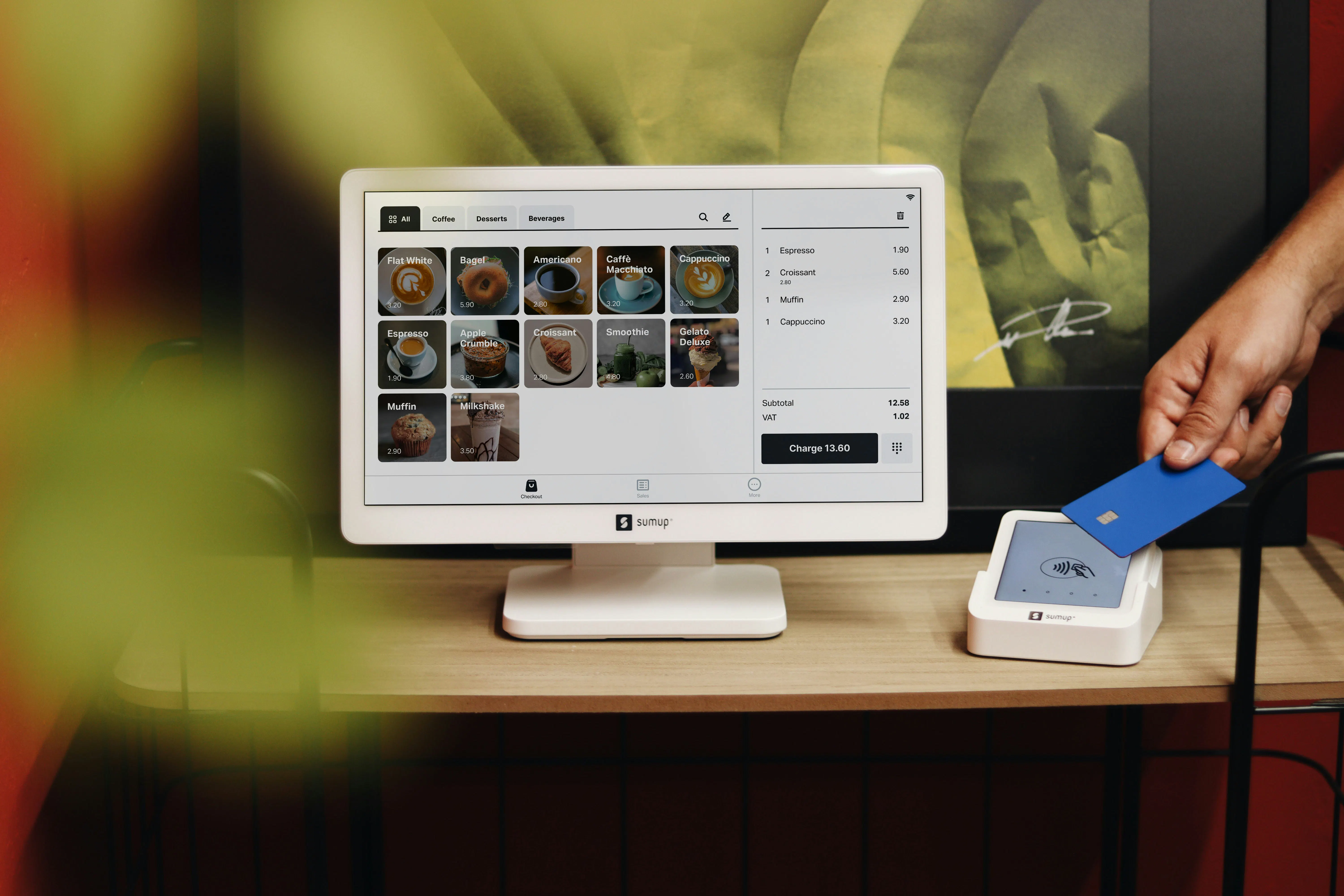A customer paying by tapping a blue credit card on a SumUp card reader next to a POS touchscreen register displaying a café menu.