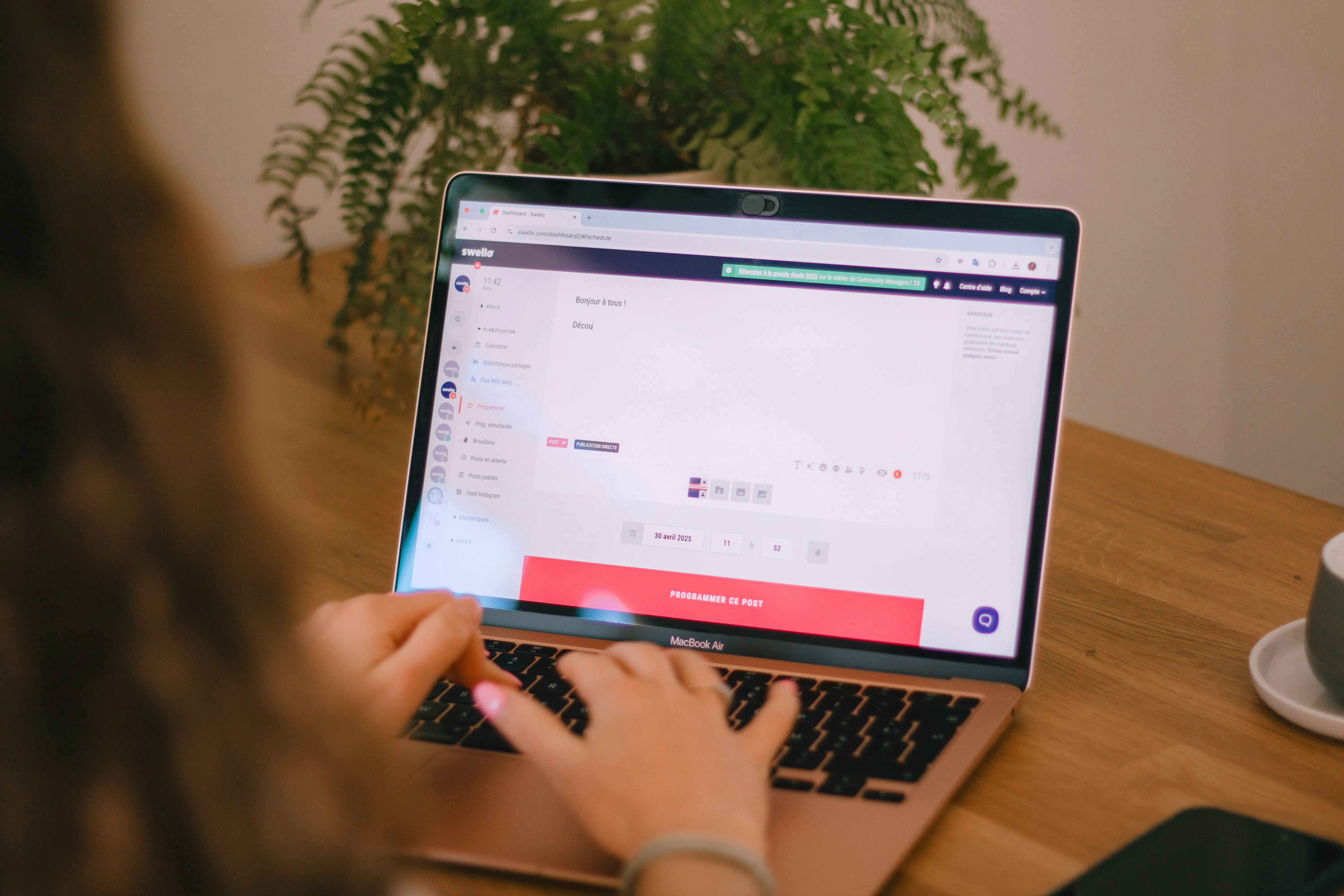 A close-up view of a person typing on a laptop keyboard, scheduling a social media post on the Swello platform at a wooden desk with a potted fern in the background