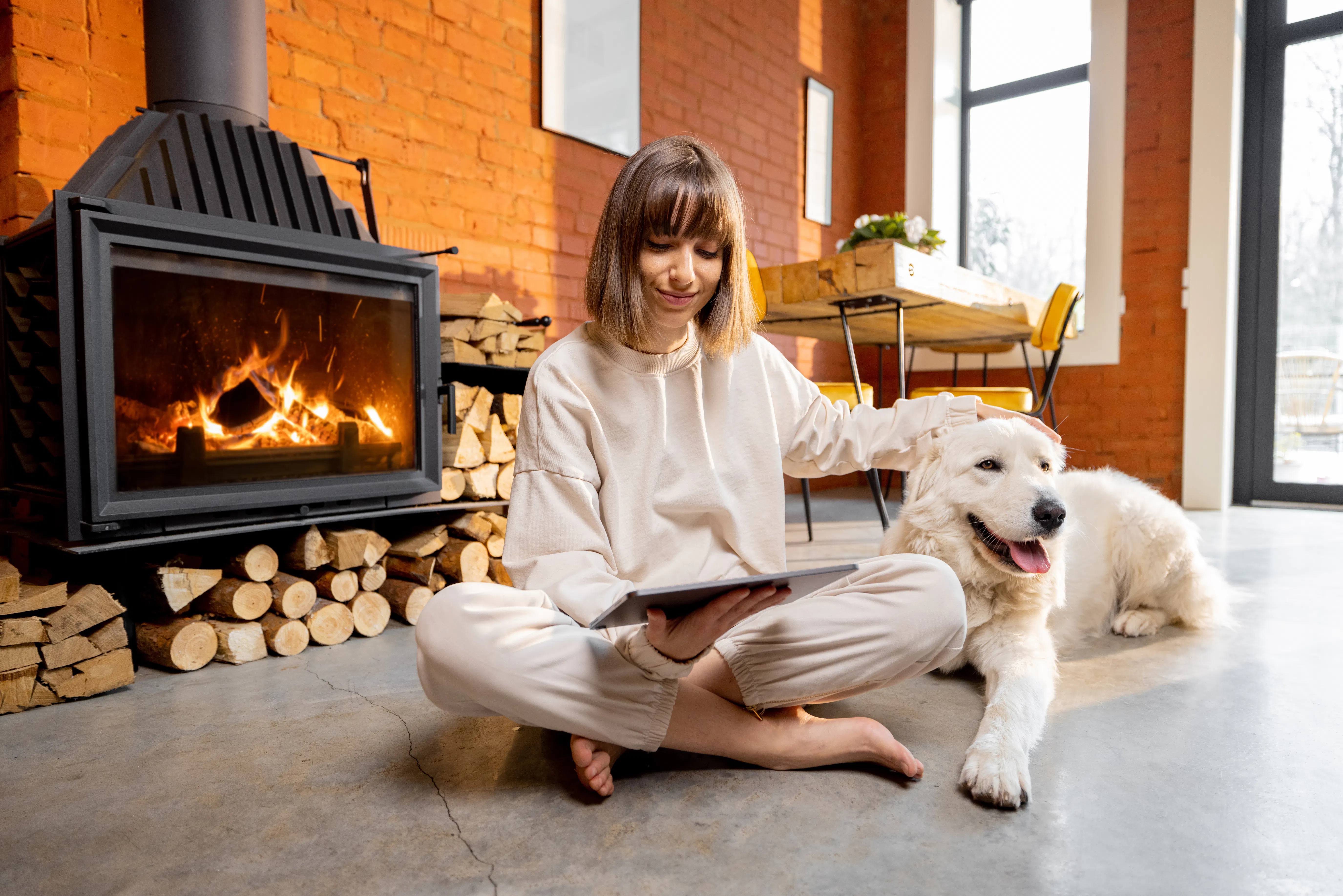Young woman sitting relaxed with her white dog on the floor near the fireplace and using digital tablet.