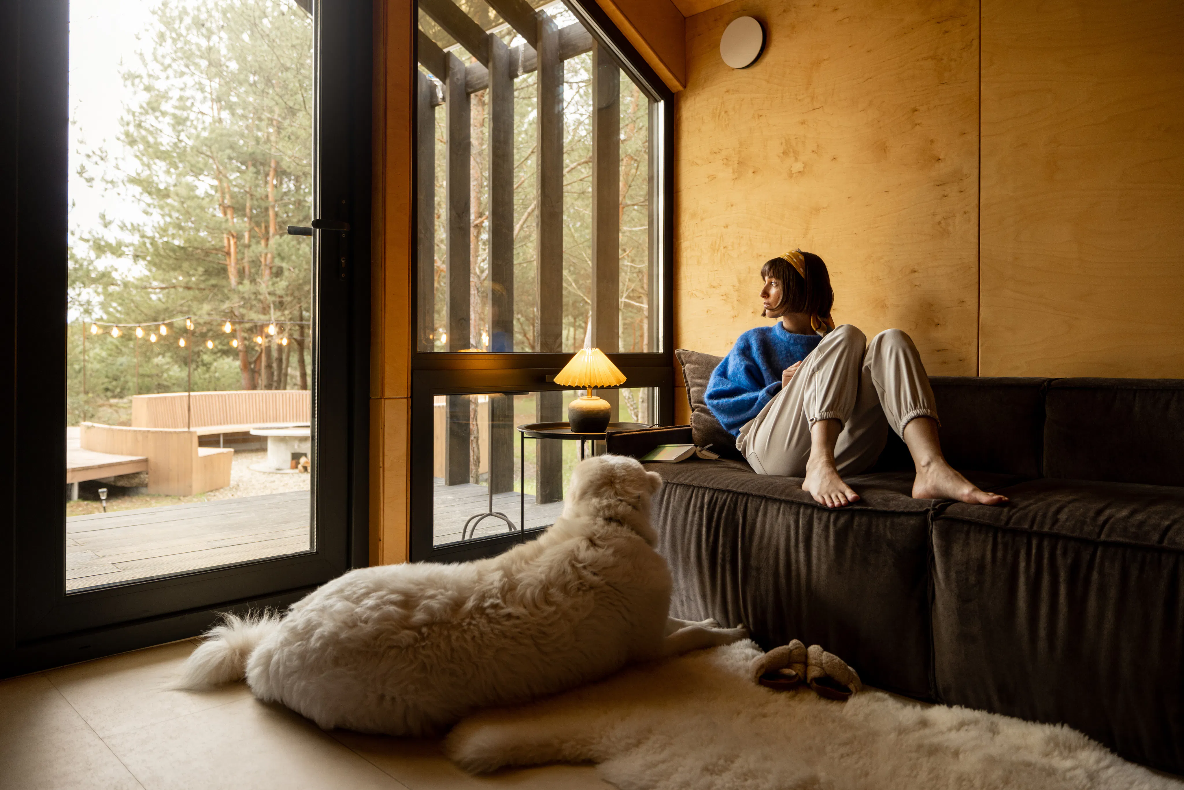 Woman sitting relaxed on a couch by the window with her dog at wooden cottage in forest