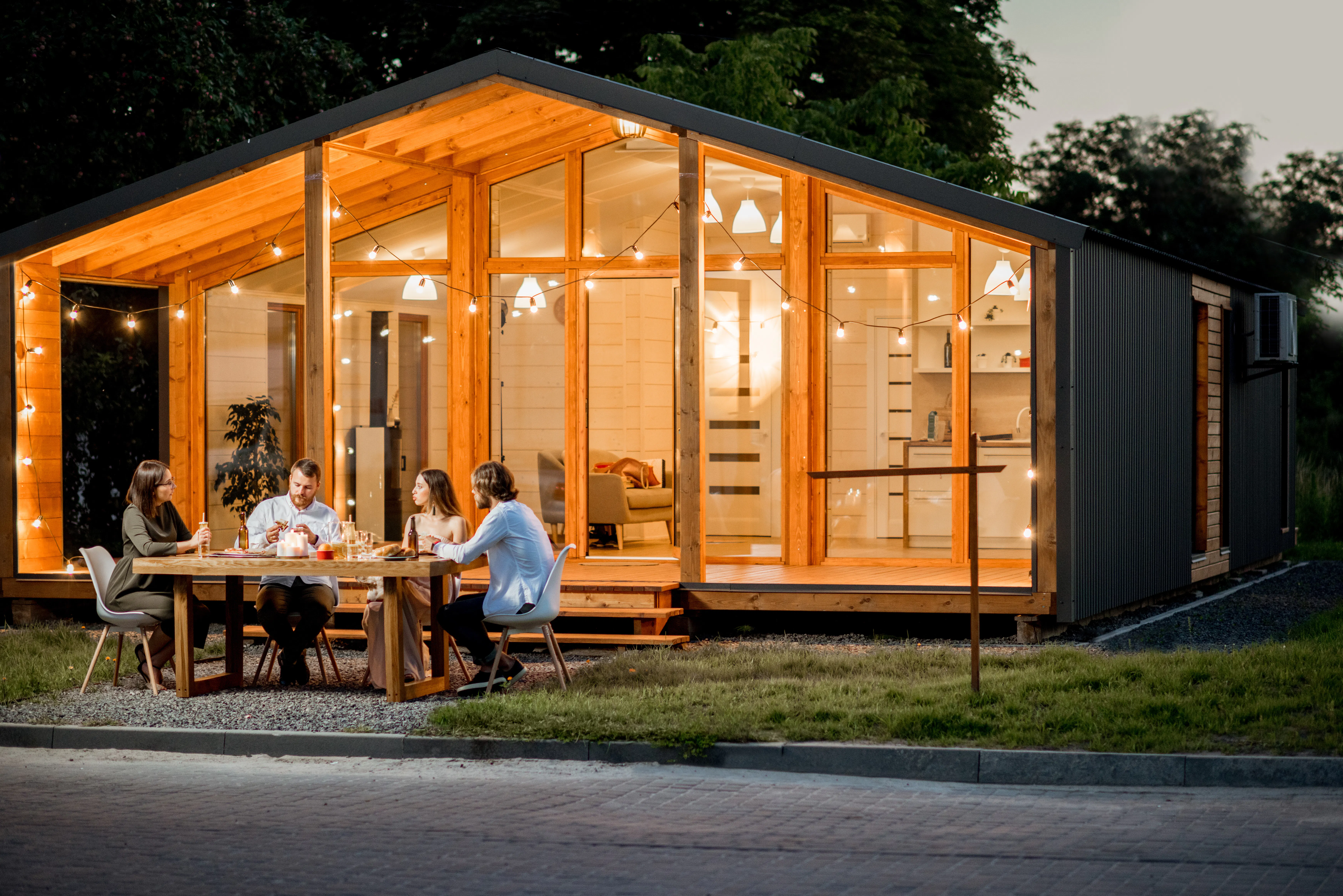 Backyard of the modern wooden house decorated with lights with friends dining at the table at the evening