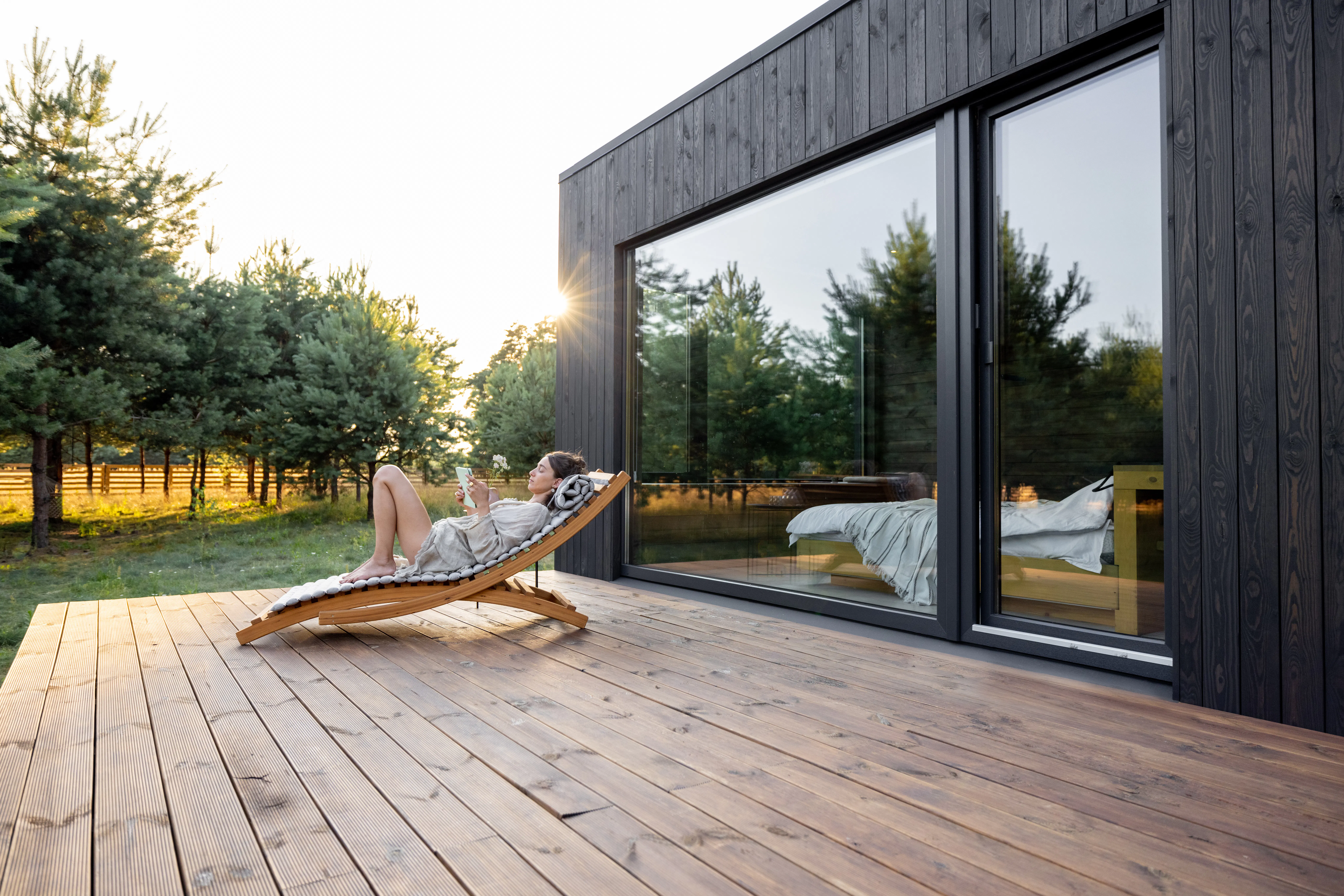 Young woman resting on sunbed and reading on a tablet on the wooden terrace near the modern house with panoramic windows near pine forest