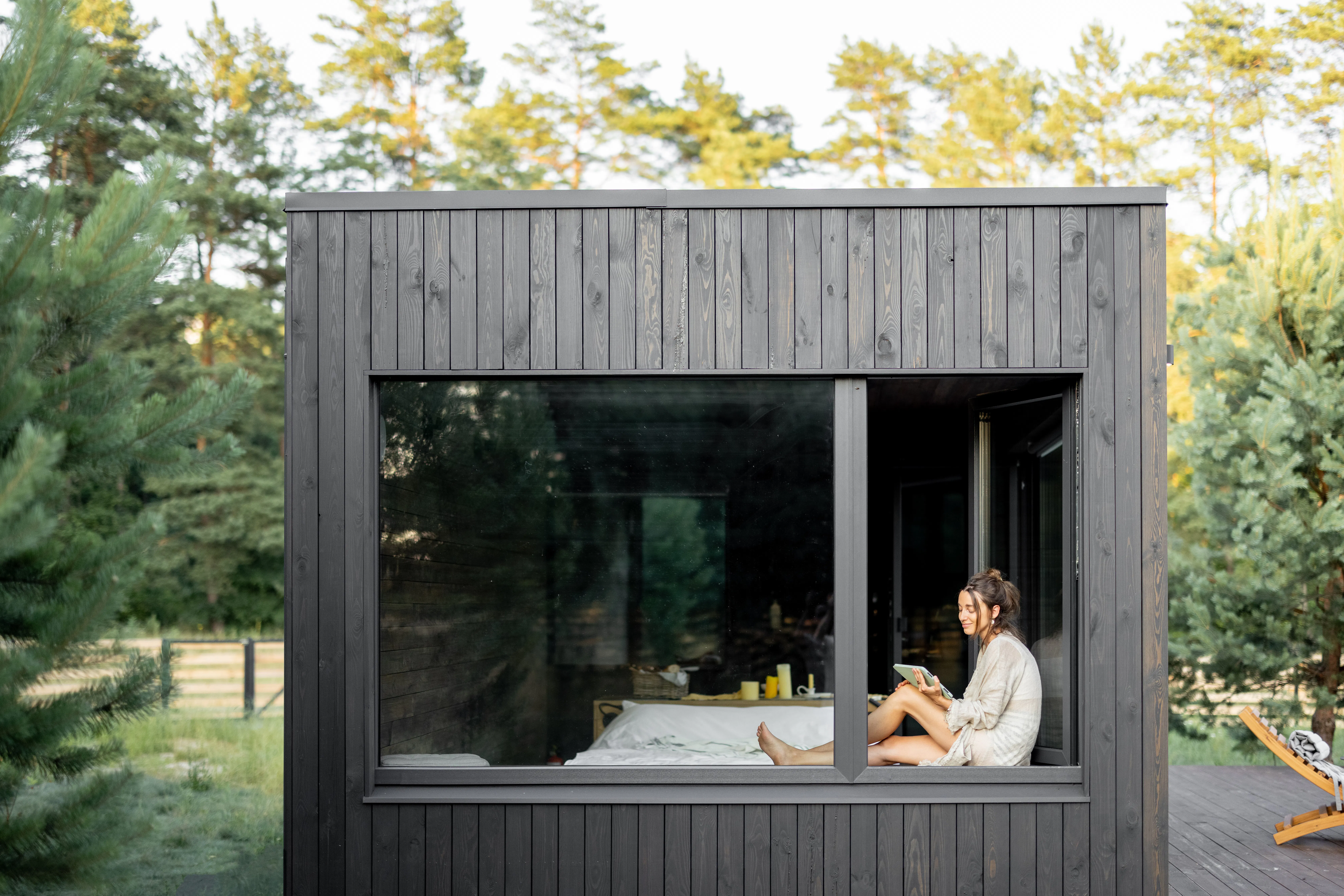 Young woman resting at beautiful country house or hotel, sitting with tablet on the window sill enjoying beautiful view on pine forest