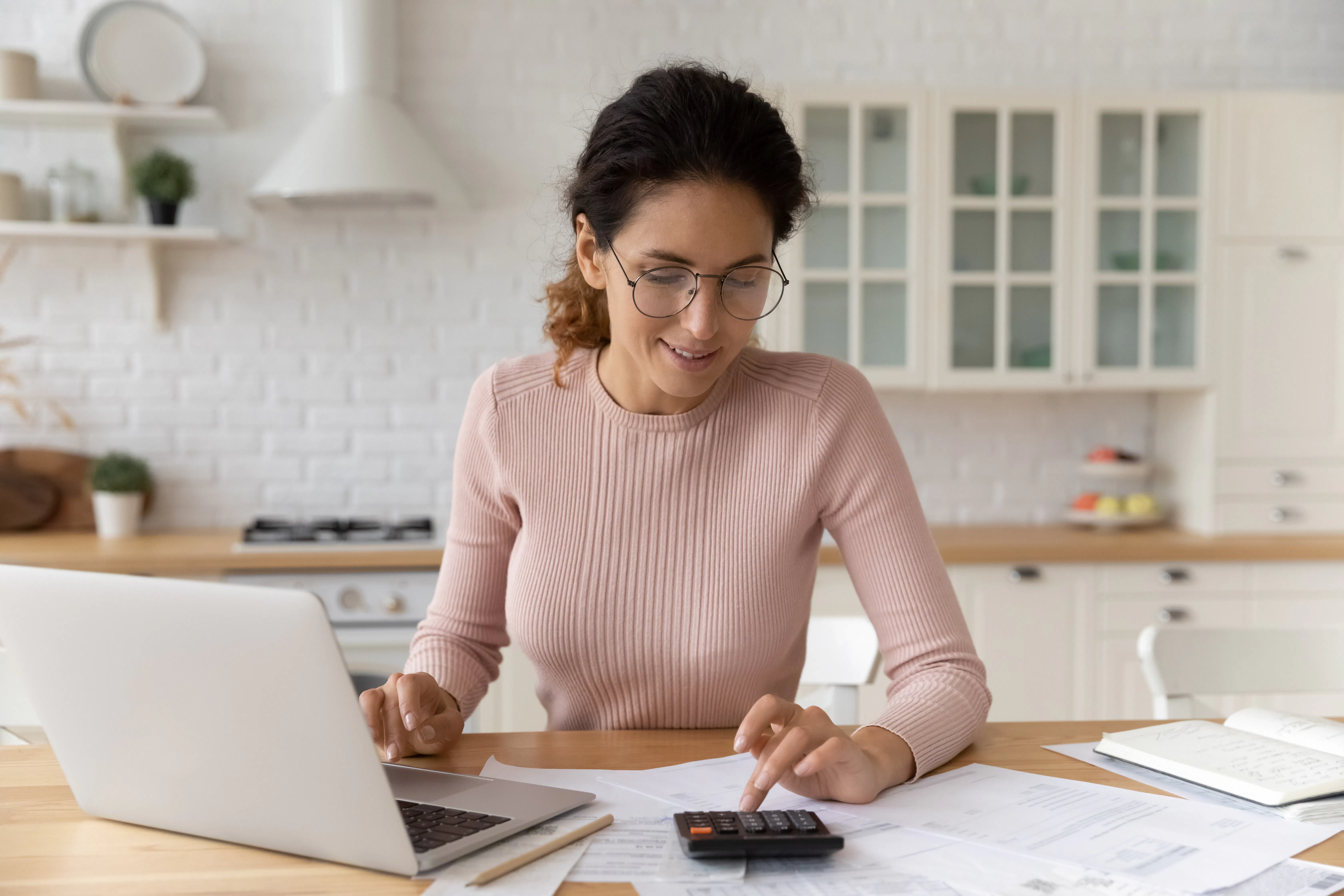 Hispanic woman use calculator calculate costs working sit at table in kitchen. Pay monthly bills through e-bank app on laptop. Business, summarize household expenses, manage budget, accounting.jpeg