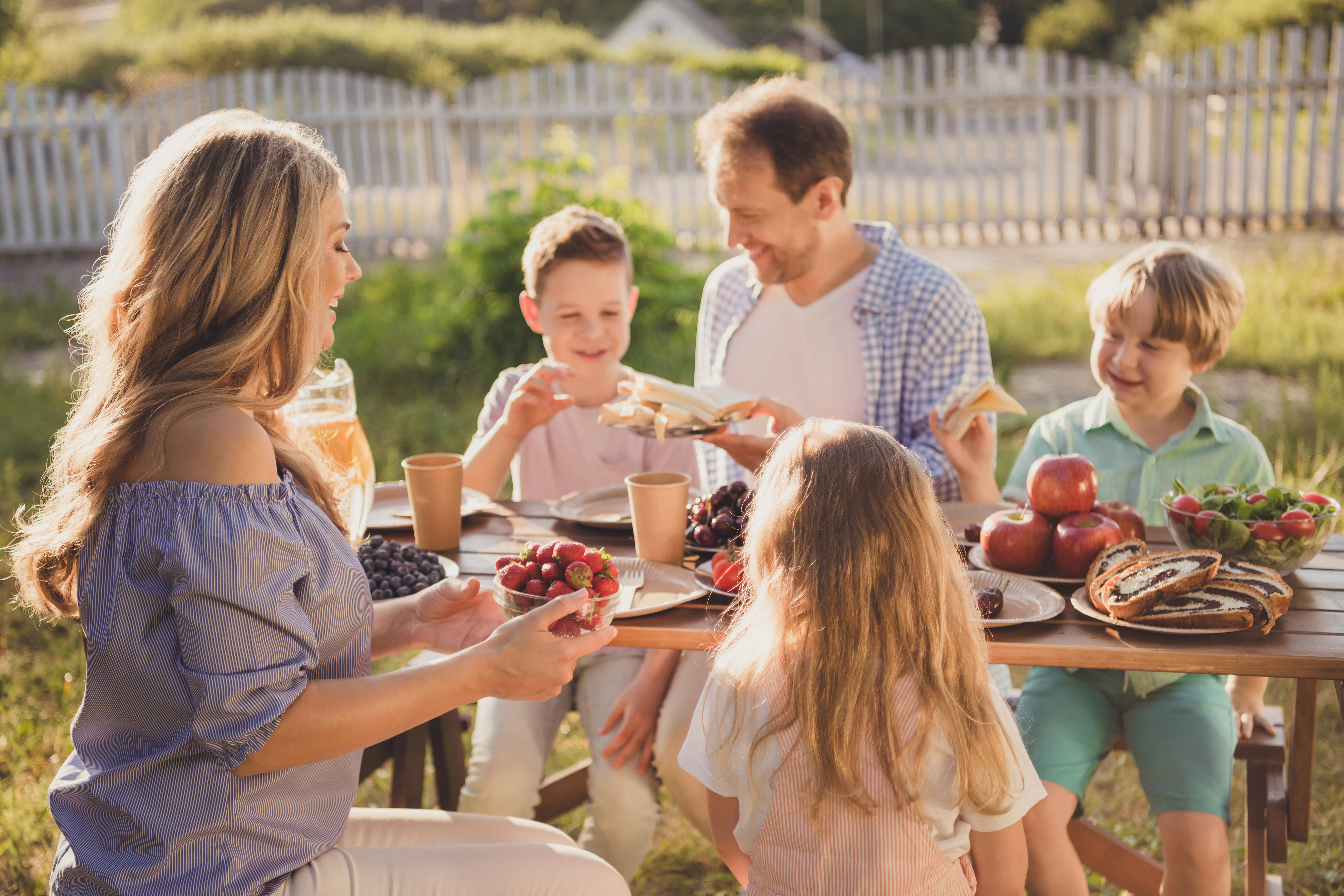Positive family people mom dad little kids children sit table enjoy summer outside countryside meal daddy share plate sandwiches son mom hold bold strawberries rest relax