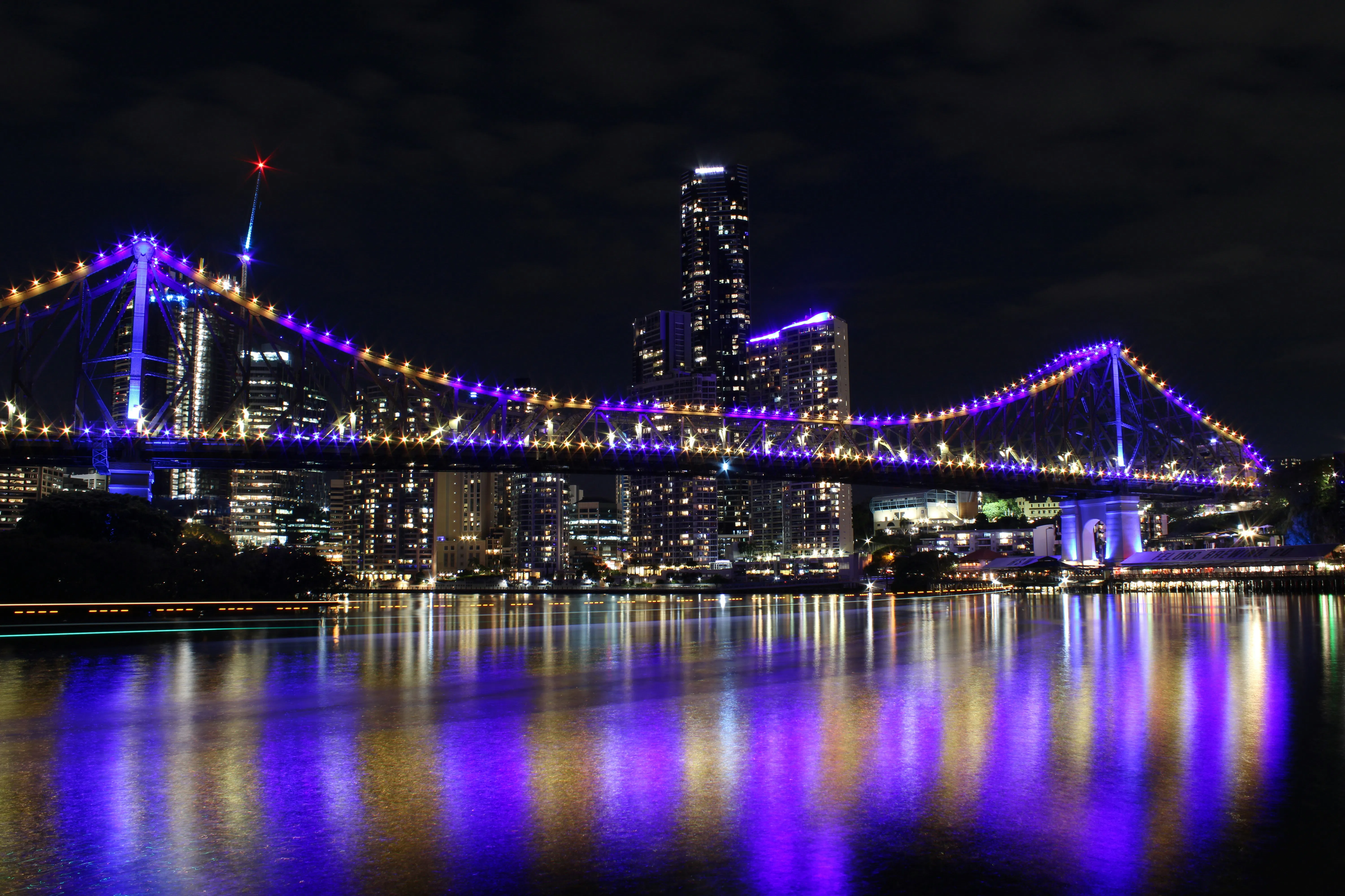 Brisbane’s Story Bridge lit in purple at night, reflecting on the river with city skyline in the background.