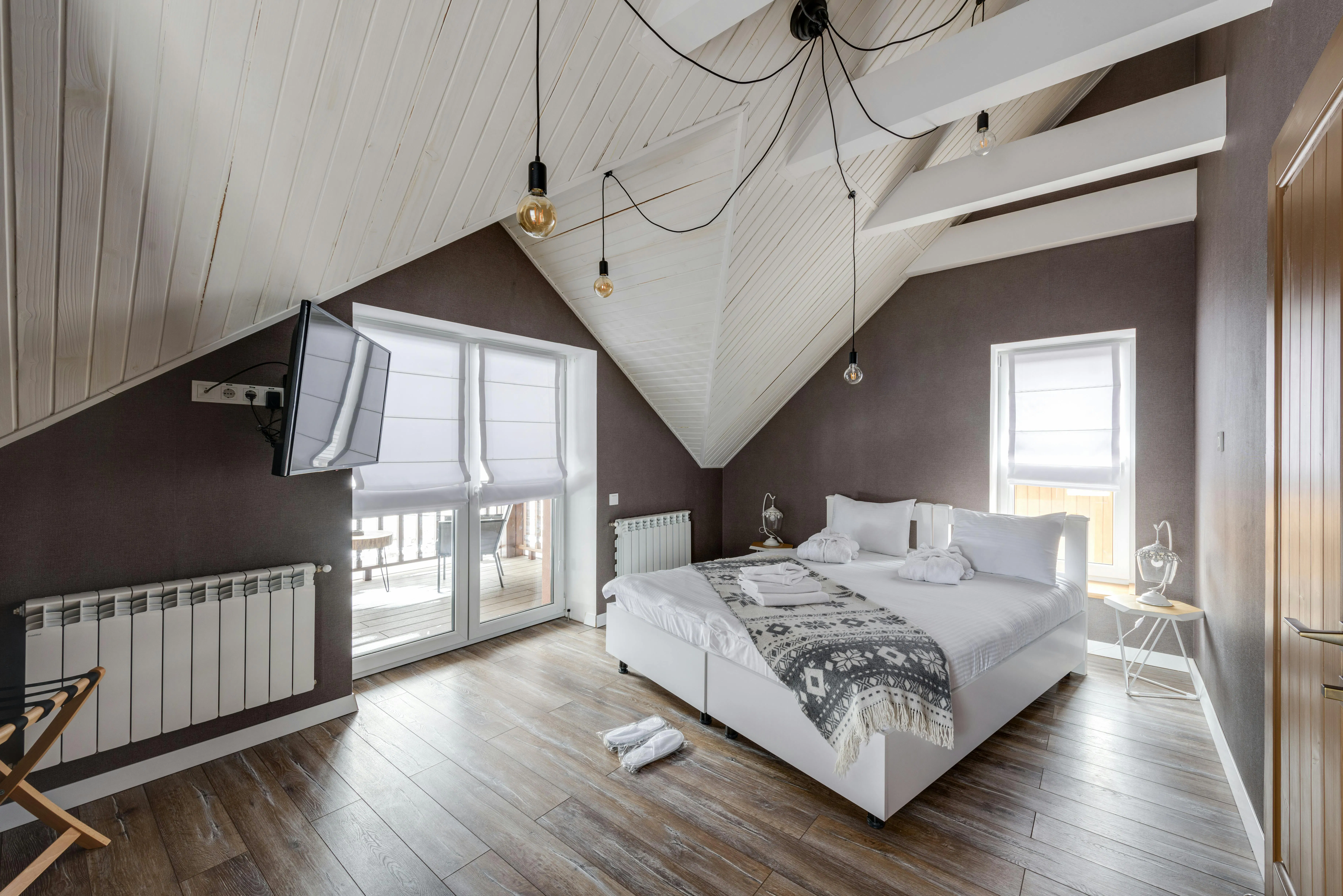 Modern attic bedroom with sloped ceiling, double bed, hanging lights, and balcony access through glass doors.