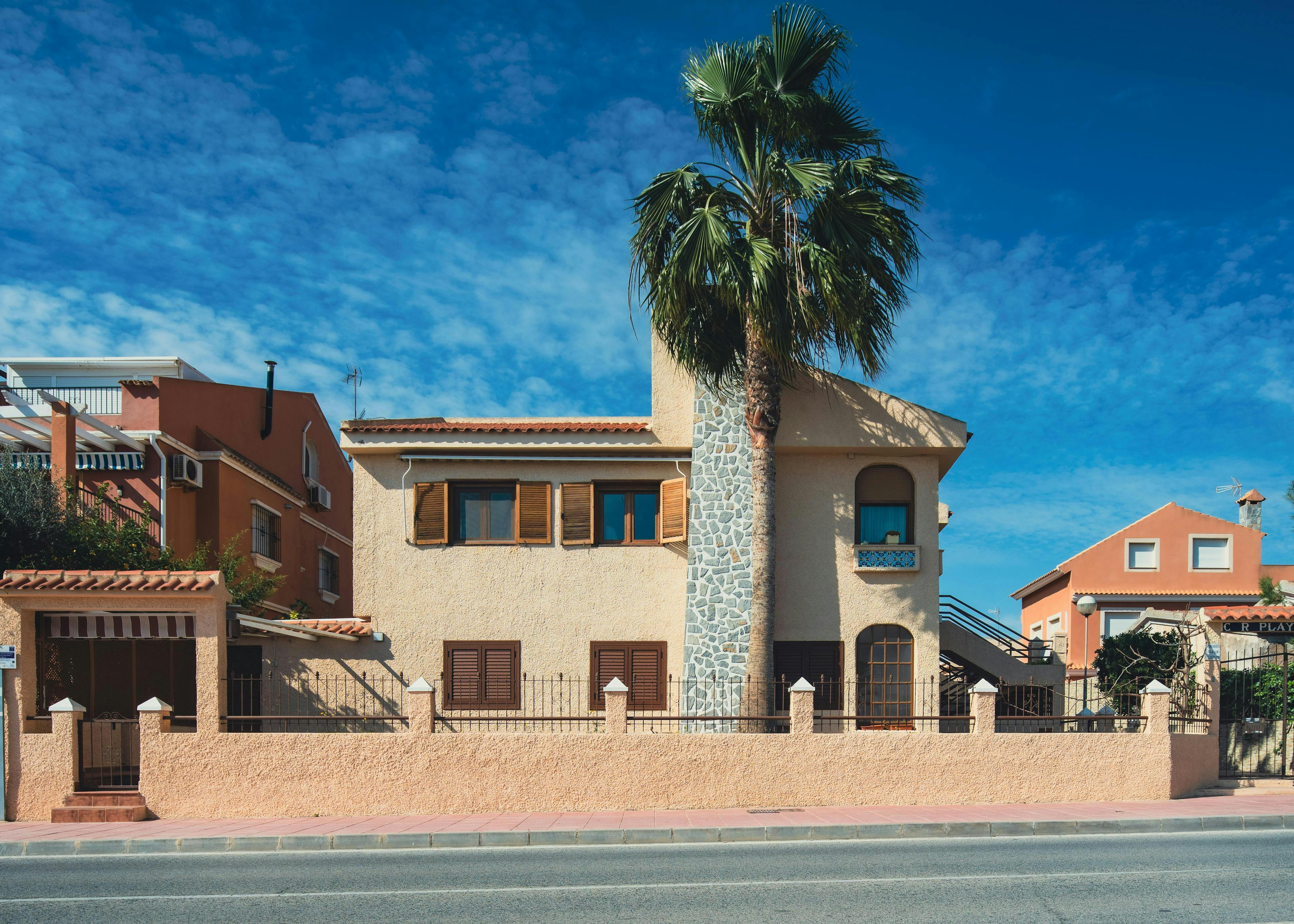 Street view of a stucco house with stone chimney and palm tree.