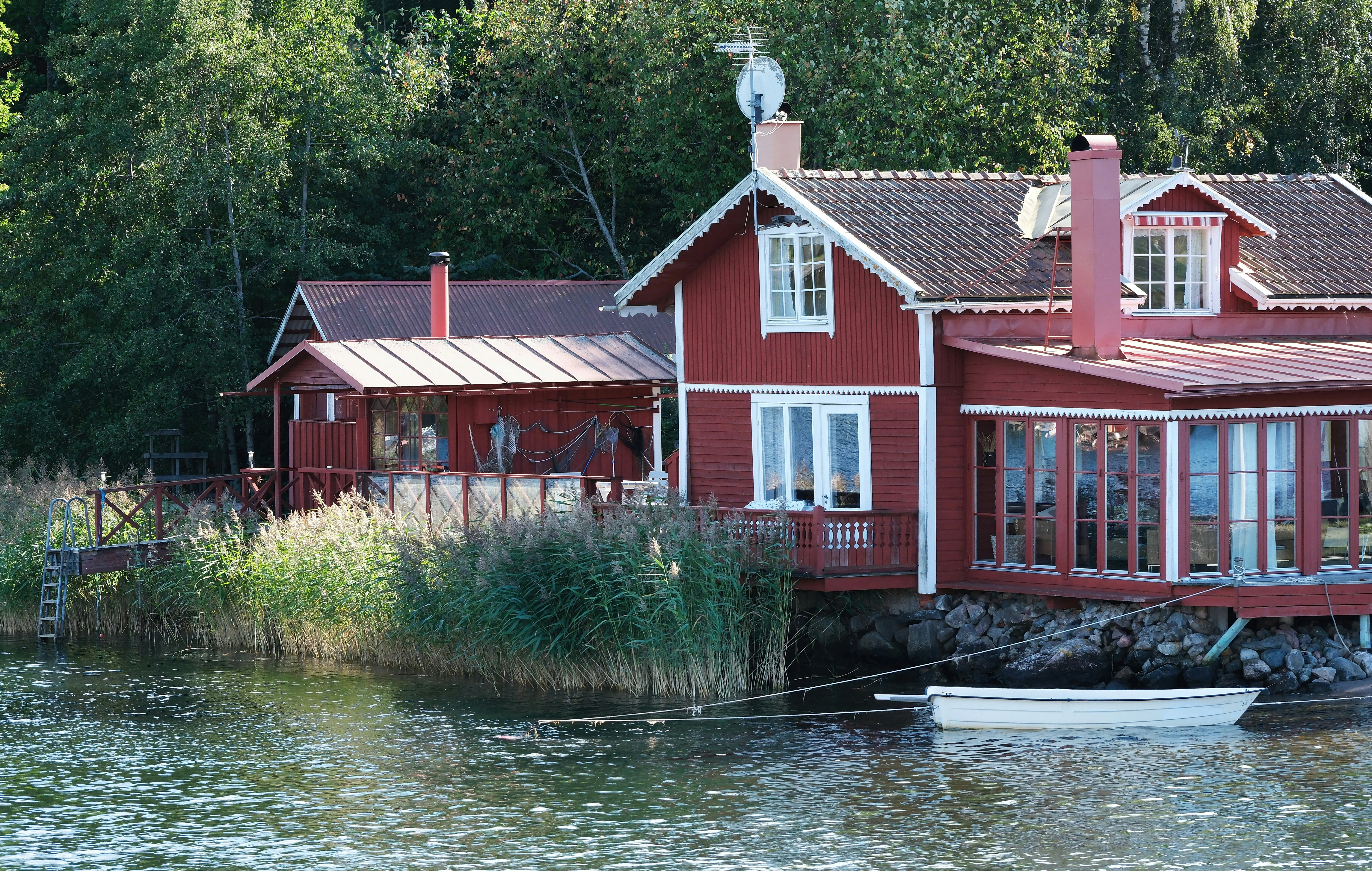 A traditional red wooden cottage on a rocky waterfront, with tall reeds and a small white rowboat.