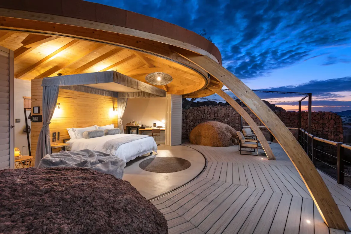Unique bedroom opening onto a circular wooden deck with large rocks and a dramatic desert sunset sky.