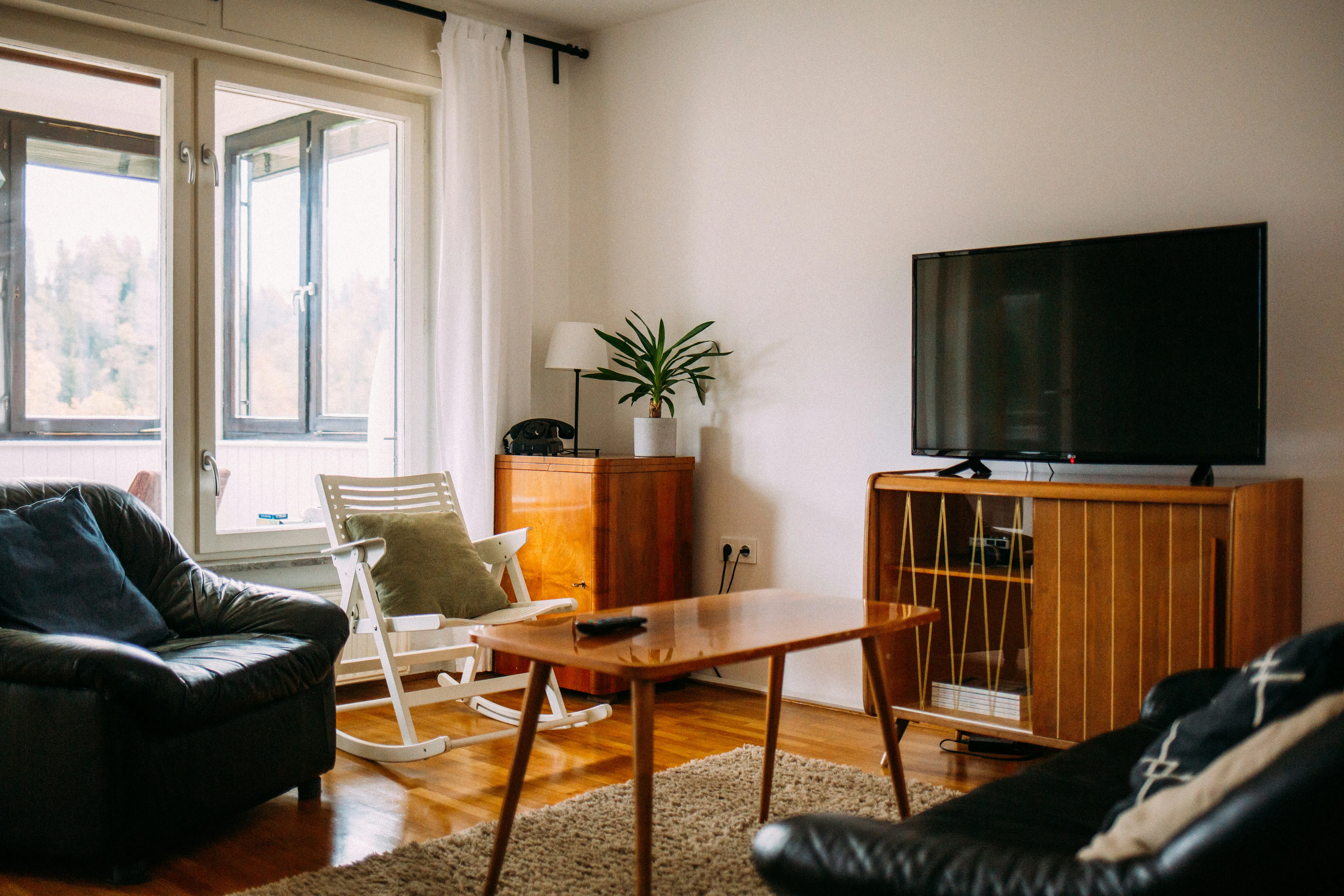 A cozy living room with mid-century modern wood furniture, a flat-screen TV, and large bright windows.
