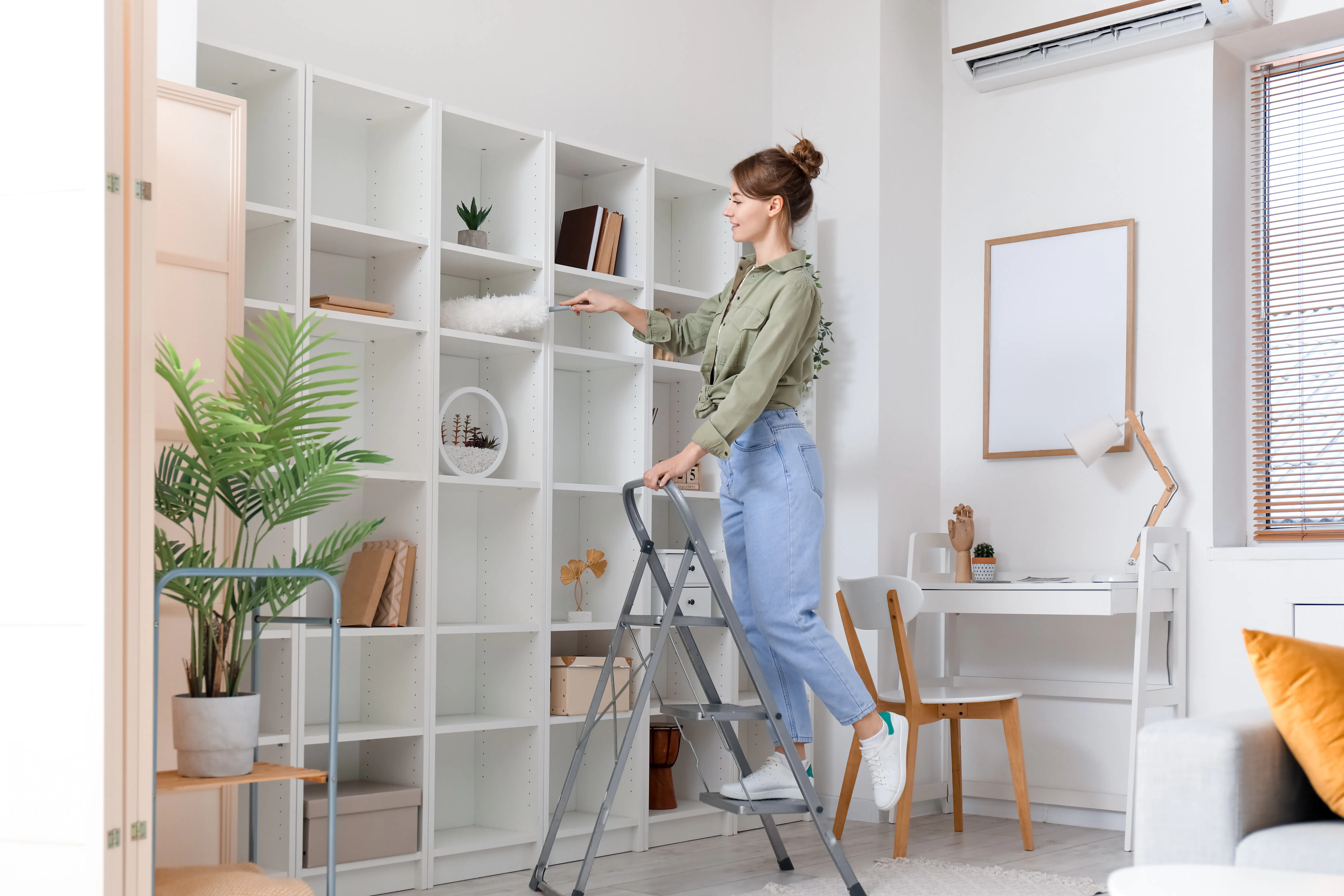 Young woman on stepladder cleaning bookshelf with duster at home