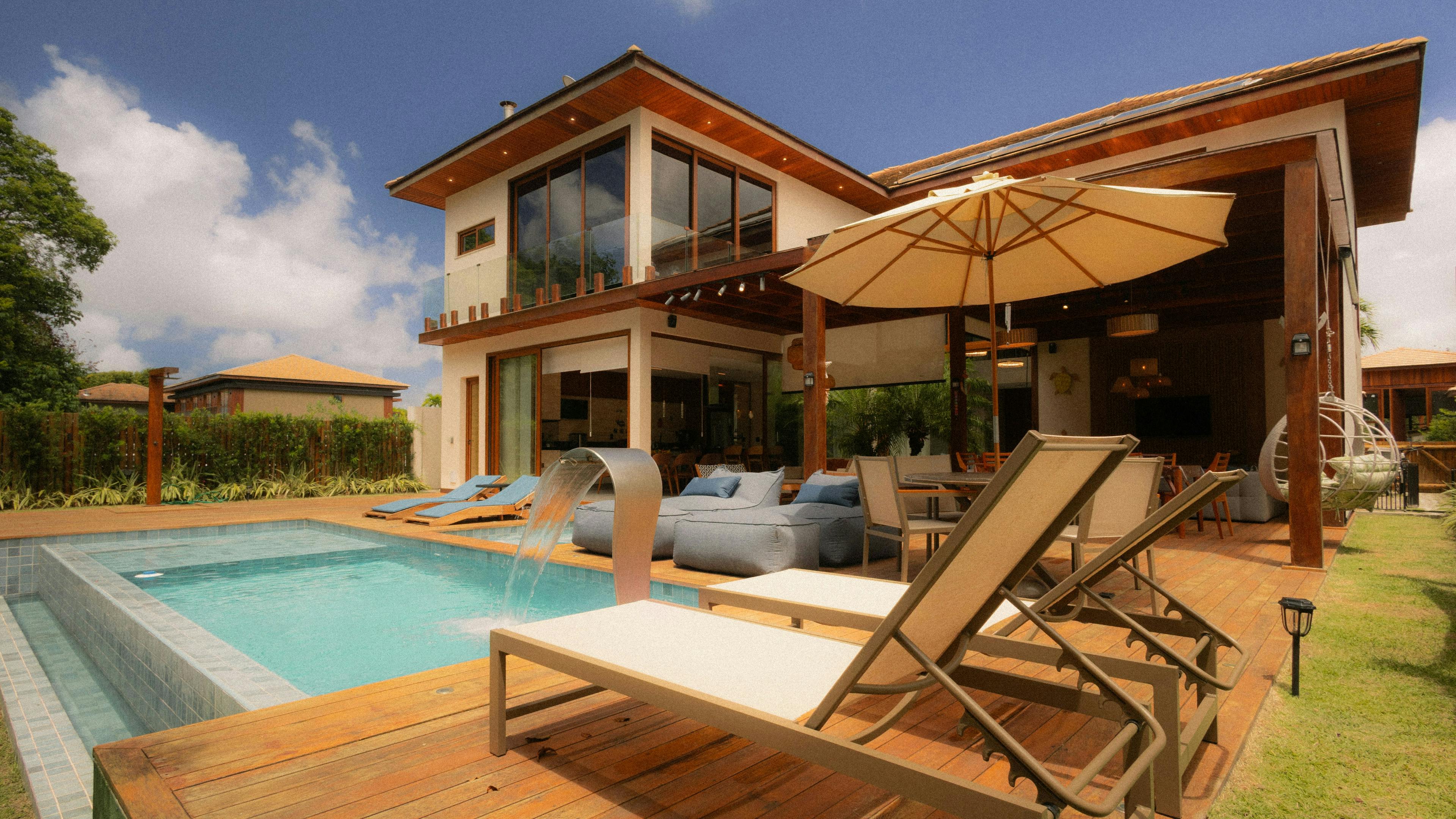 Modern two-story vacation home with a wooden deck, lounge chairs, a pool with a waterfall feature, and a patio.