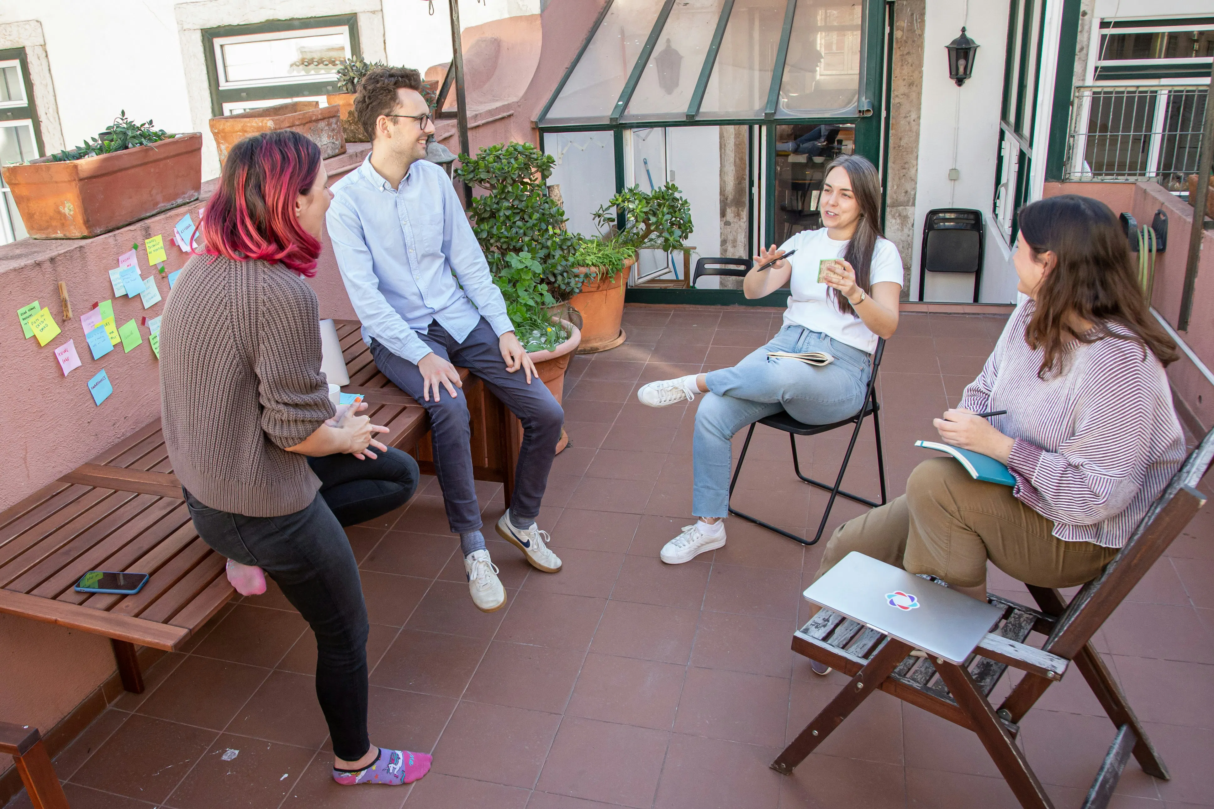 Four people sitting on a sunny rooftop terrace engaged in a meeting, with colorful sticky notes posted on a pink wall and plants in the background.