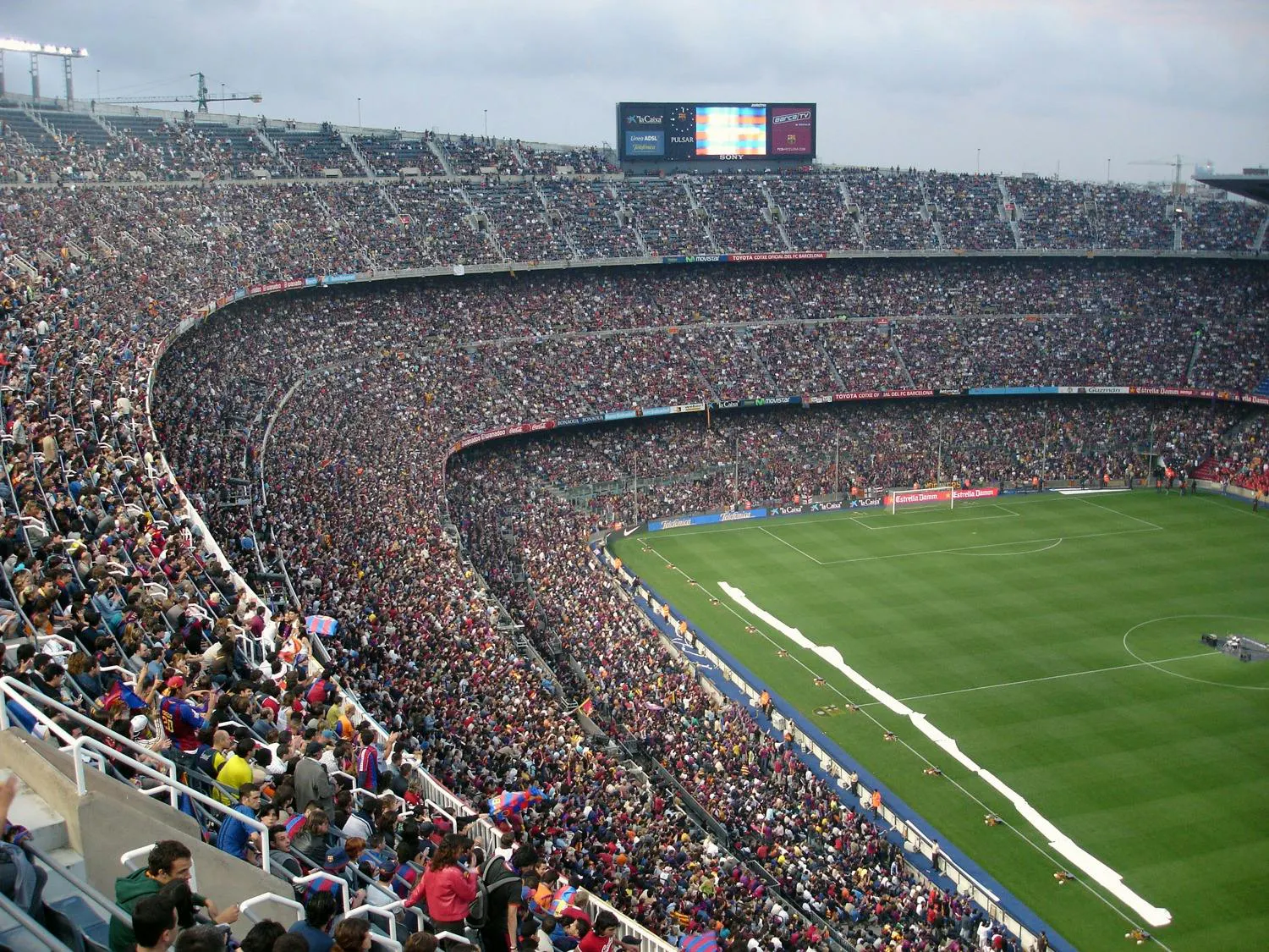 A high-angle view of a massive, crowded soccer stadium filled with thousands of fans watching a match on a bright green field.
