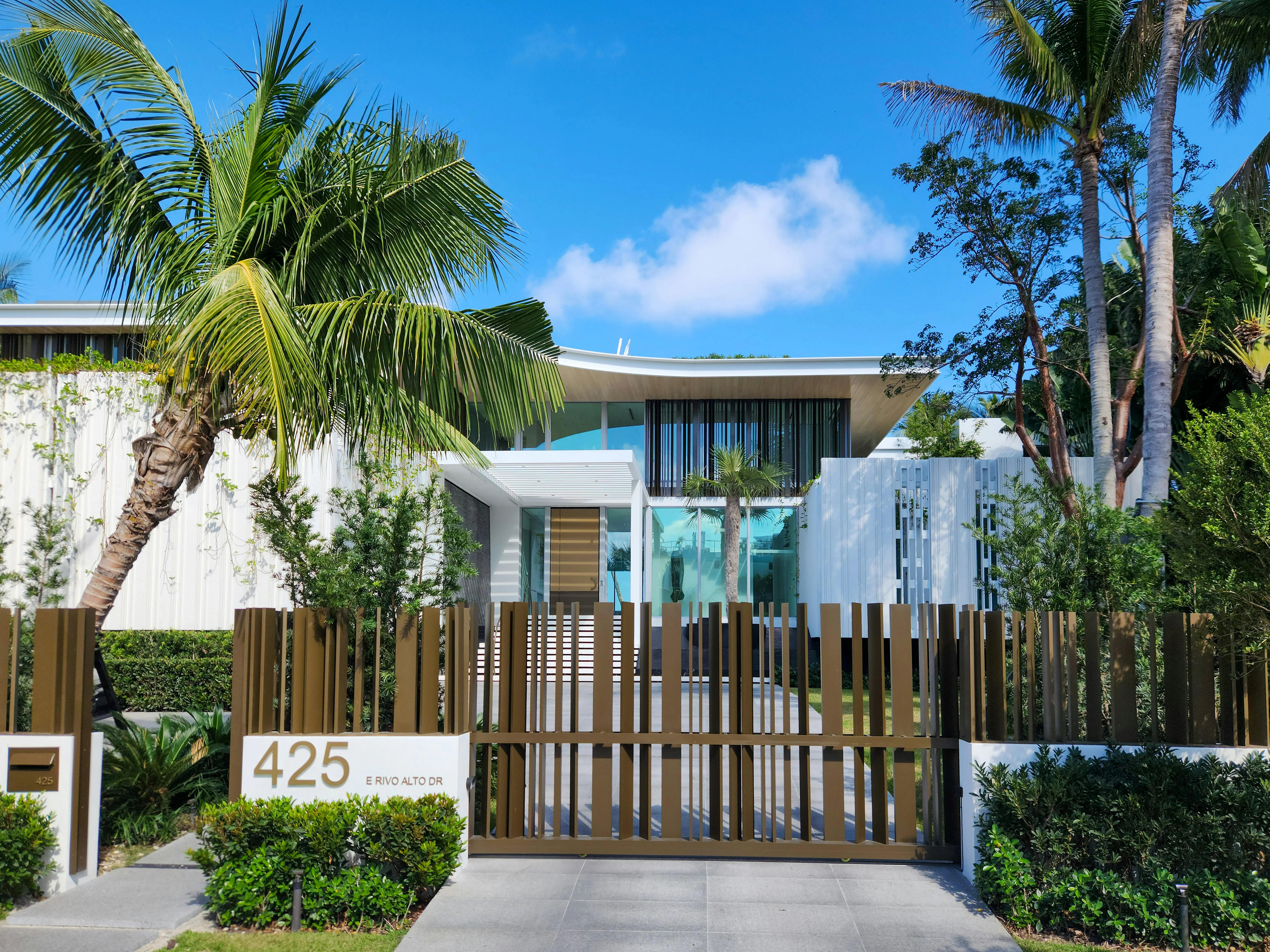 A modern white luxury home in Miami with a gated entrance and palm trees under a bright blue sky.