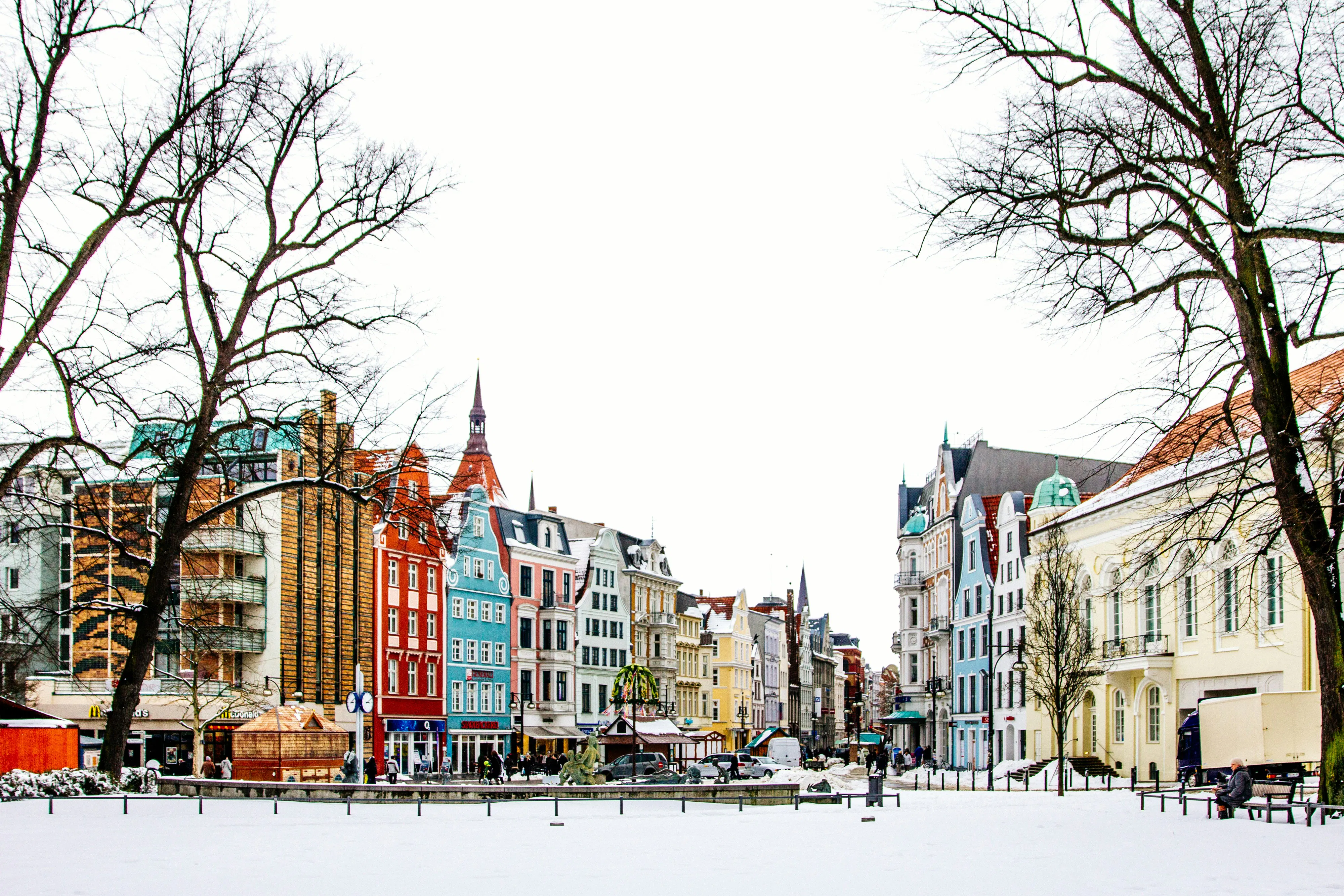 Verschneiter Universitätsplatz in Rostock mit farbenfrohen Giebelhäusern und kahlen Bäumen unter hellem Himmel.