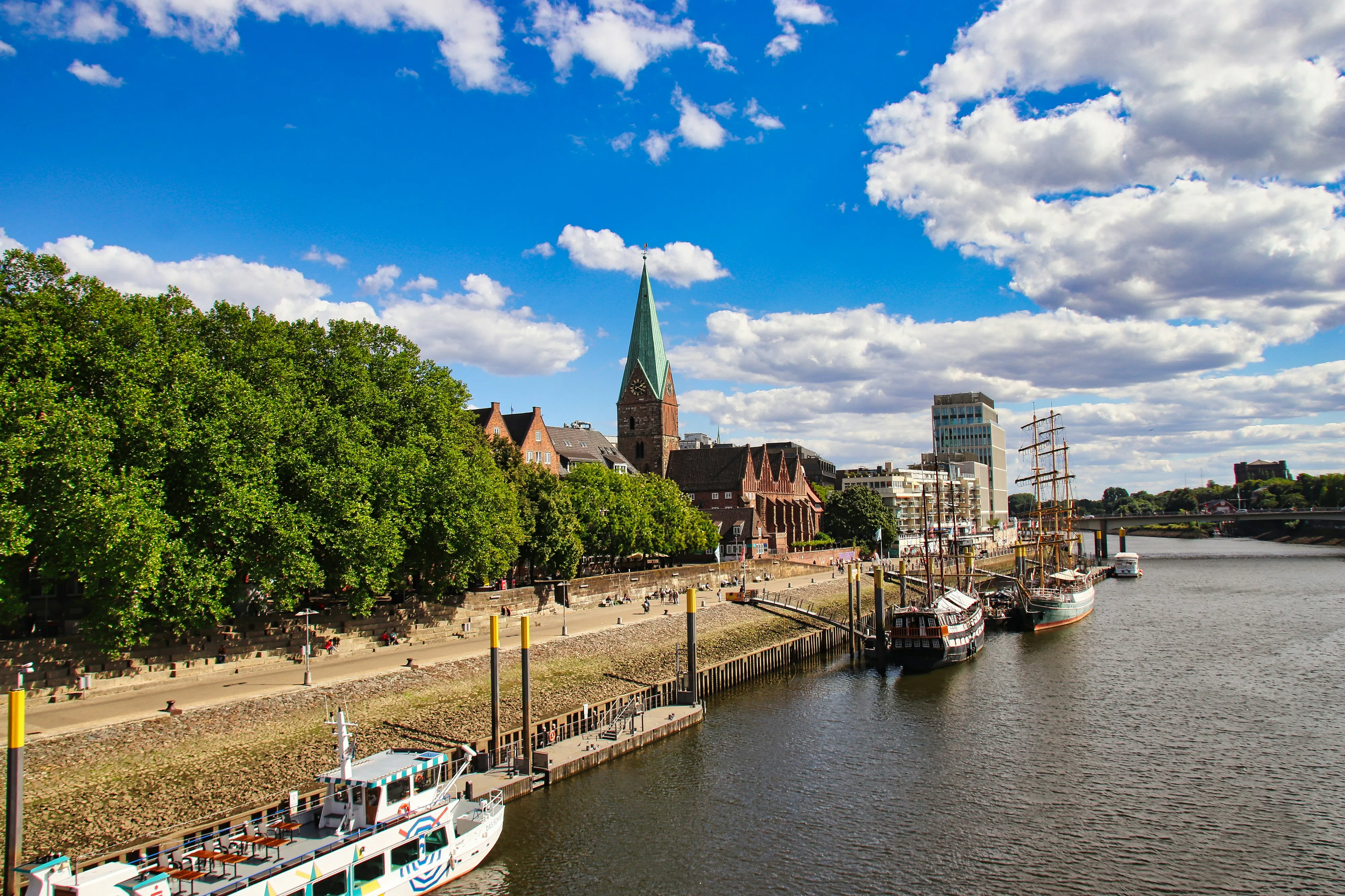 Blick auf die Uferpromenade in Bremen mit der markanten St. Martini Kirche und Segelschiffen an der Weser.