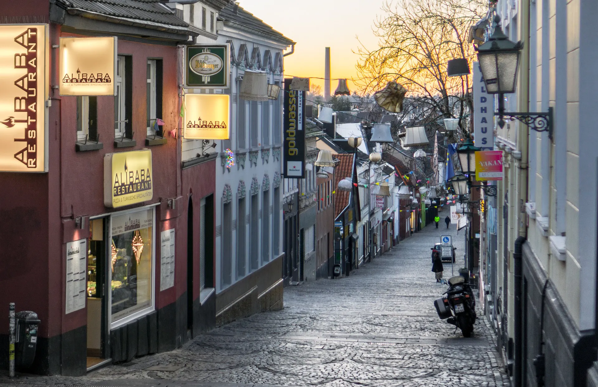 Blick in eine abschüssige Kopfsteinpflastergasse in Mönchengladbach mit Restaurants und hängenden Lampen zur Abenddämmerung.