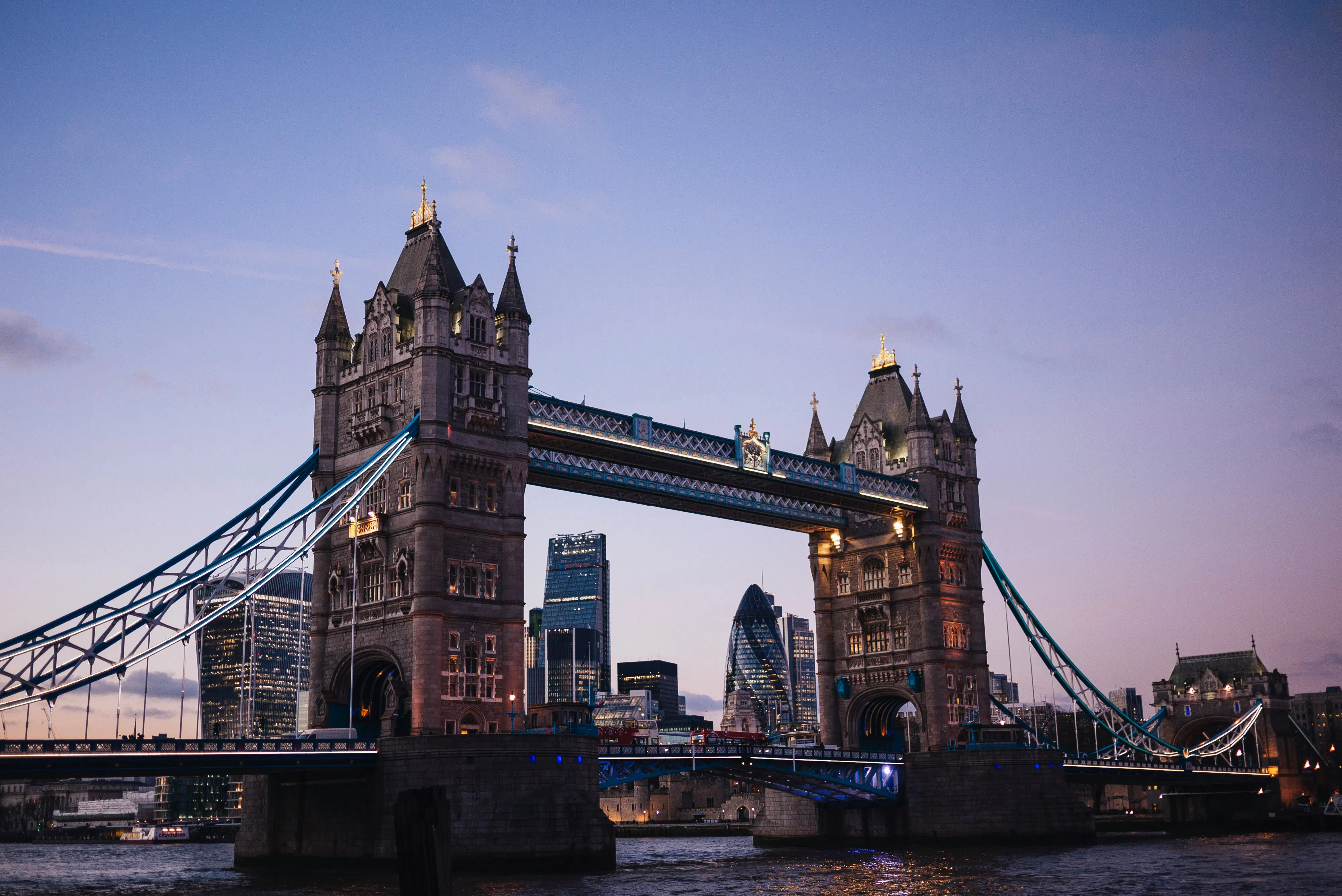 Tower Bridge illuminated at dusk with the London city skyline and skyscrapers visible in the background.