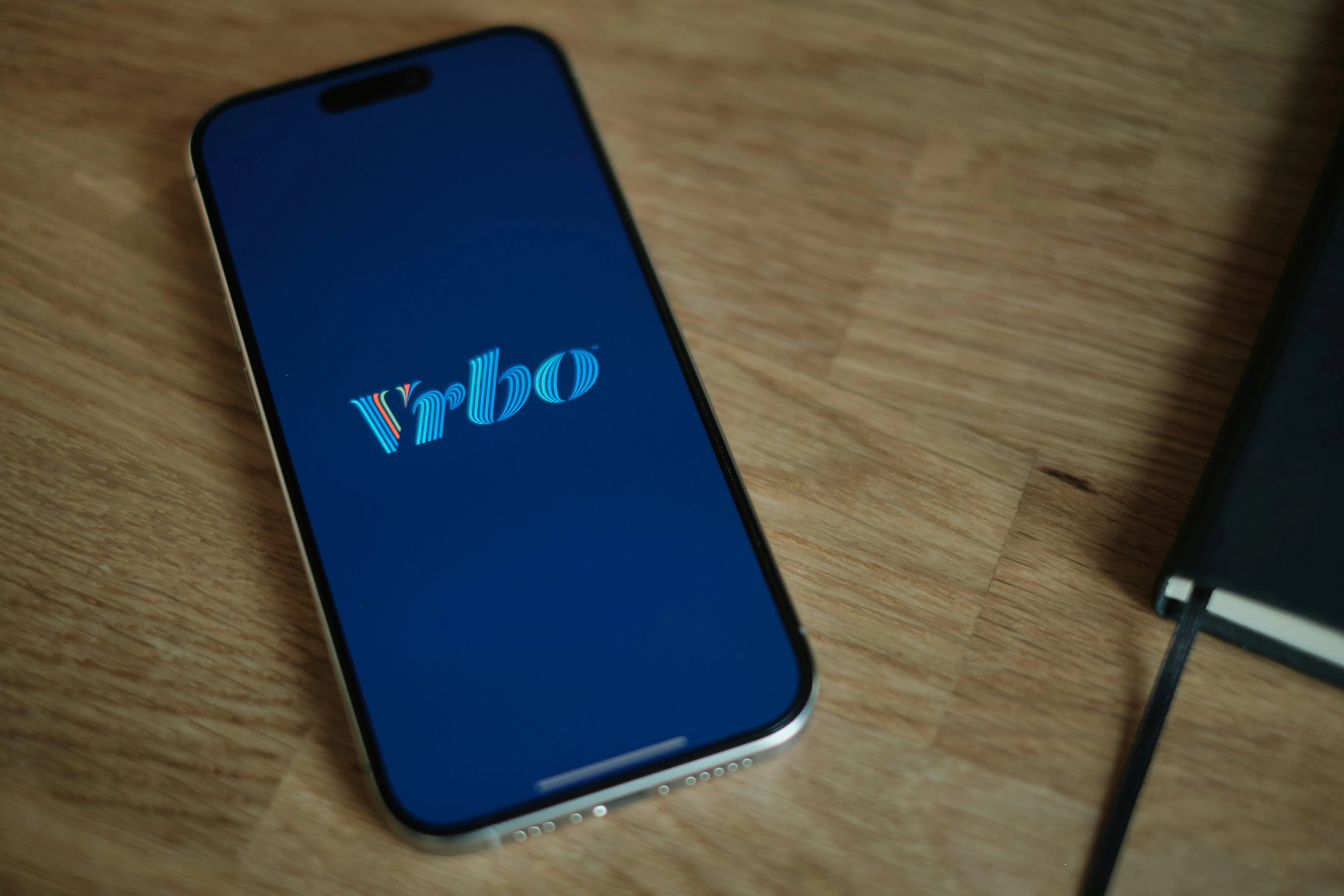 The Vrbo vacation rental app logo displayed on a smartphone screen resting on a wooden surface.