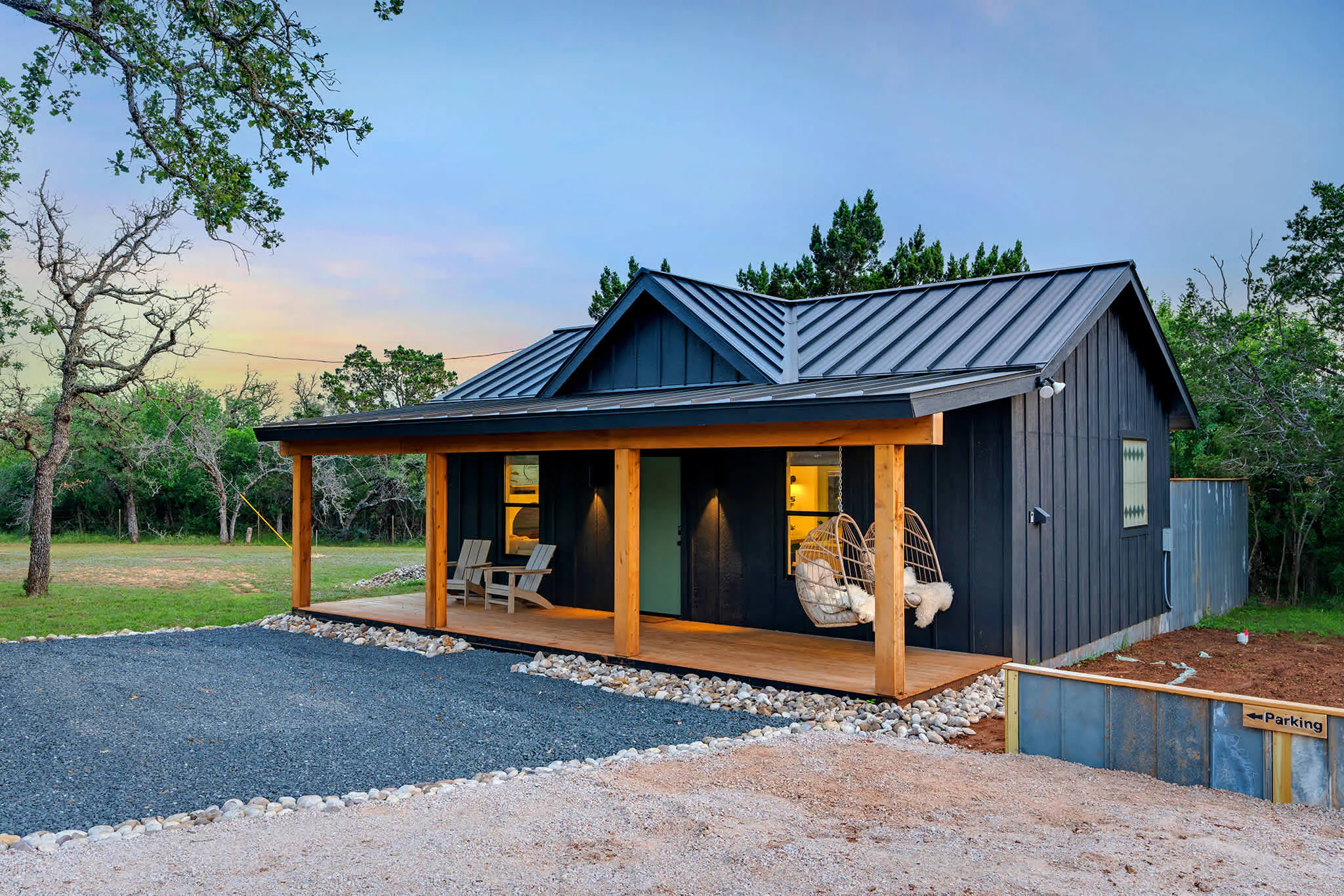Black board-and-batten cottage managed by Bach Bros Properties in the Texas Hill Country, with a cedar front porch, hanging egg chairs and a metal roof