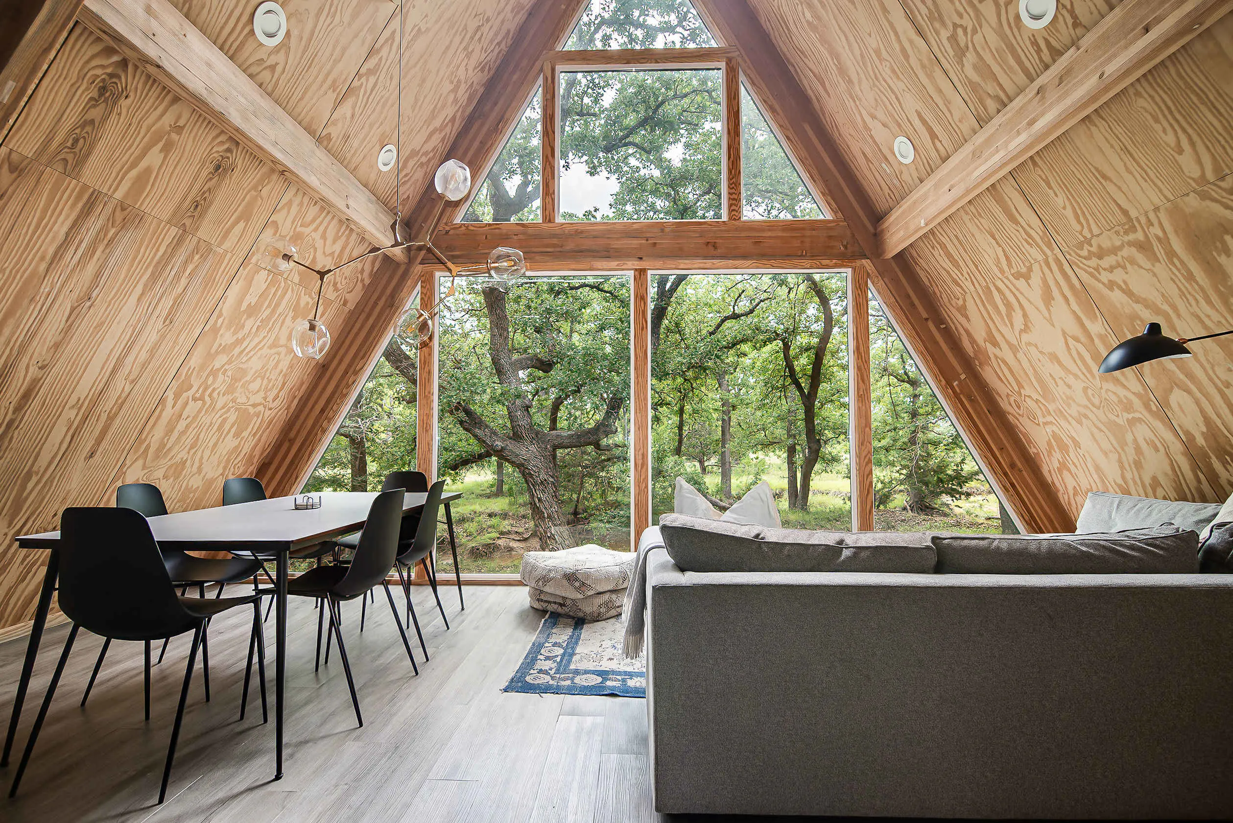 Inside a Bach Bros Properties A-frame cabin with a dining table, sectional sofa and floor-to-ceiling windows facing oak trees