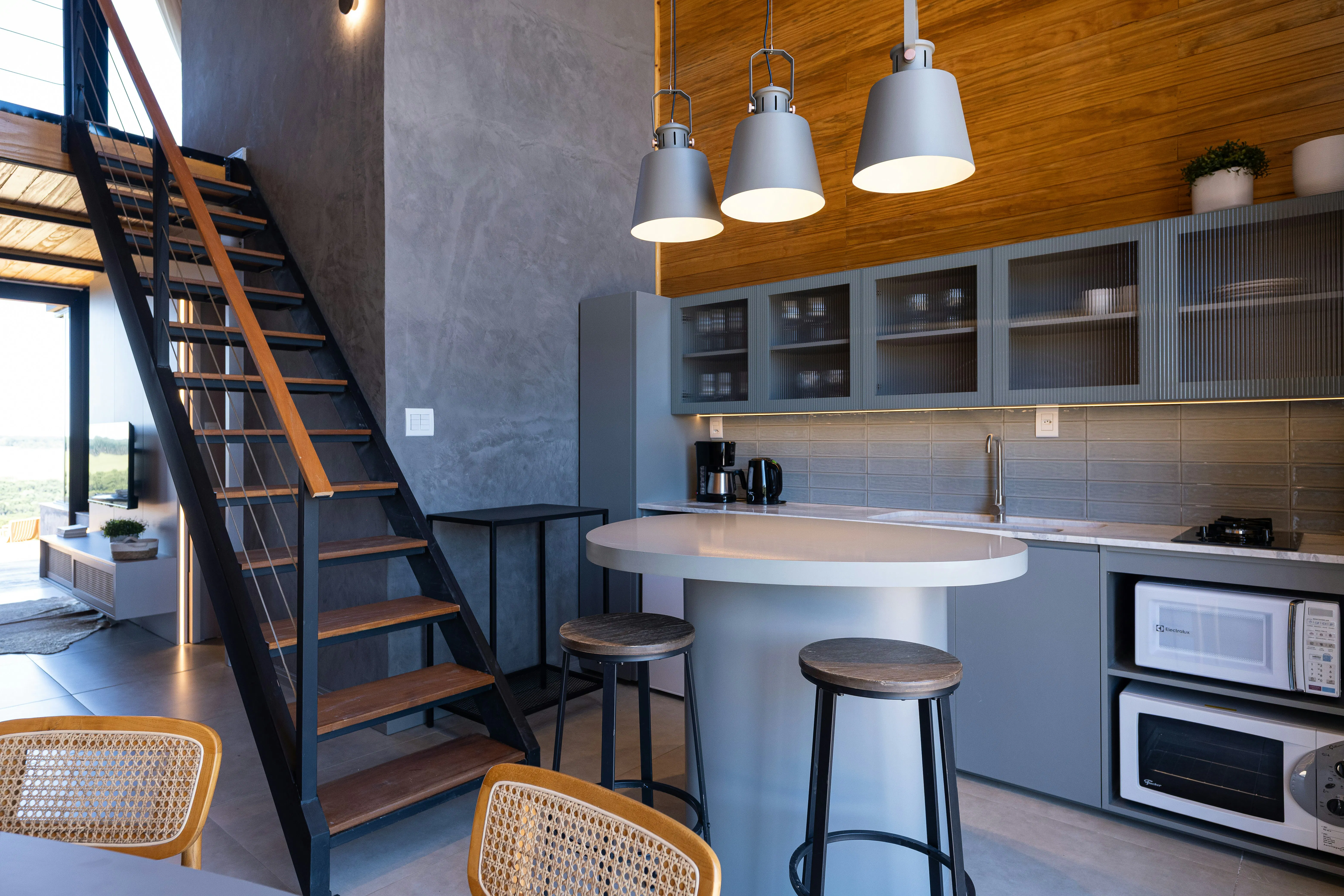 A modern kitchen in a loft featuring grey cabinets, a kitchen island with bar stools, pendant lights, a wooden accent wall, and a metal staircase