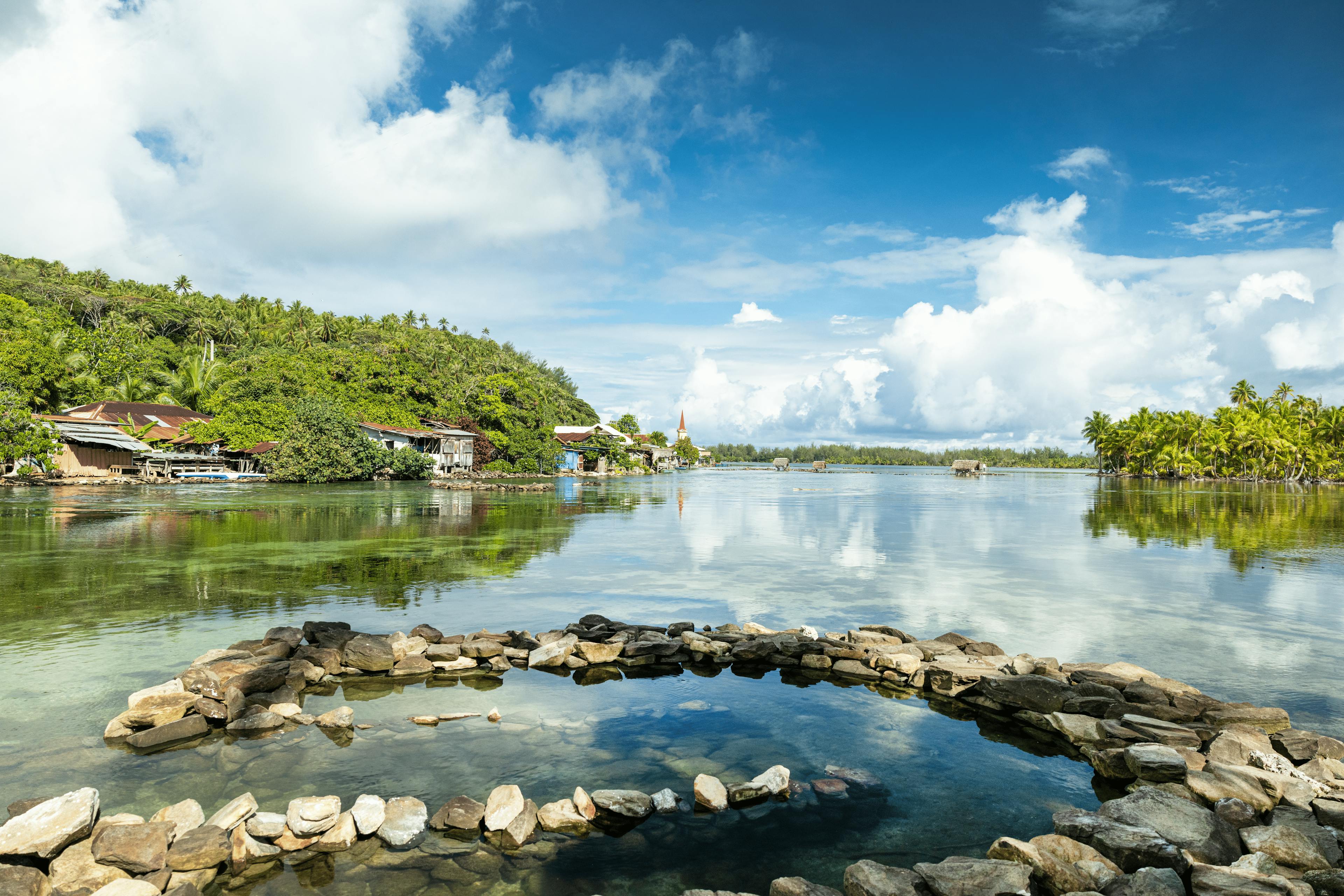 Vue idyllique d'un paisible village au bord de l'eau à Huahine, montrant des maisons traditionnelles polynésiennes nichées parmi une verdure luxuriante, avec un étang à poissons construit en roches au premier plan et un lagon calme reflétant le ciel bleu et les nuages duveteux.