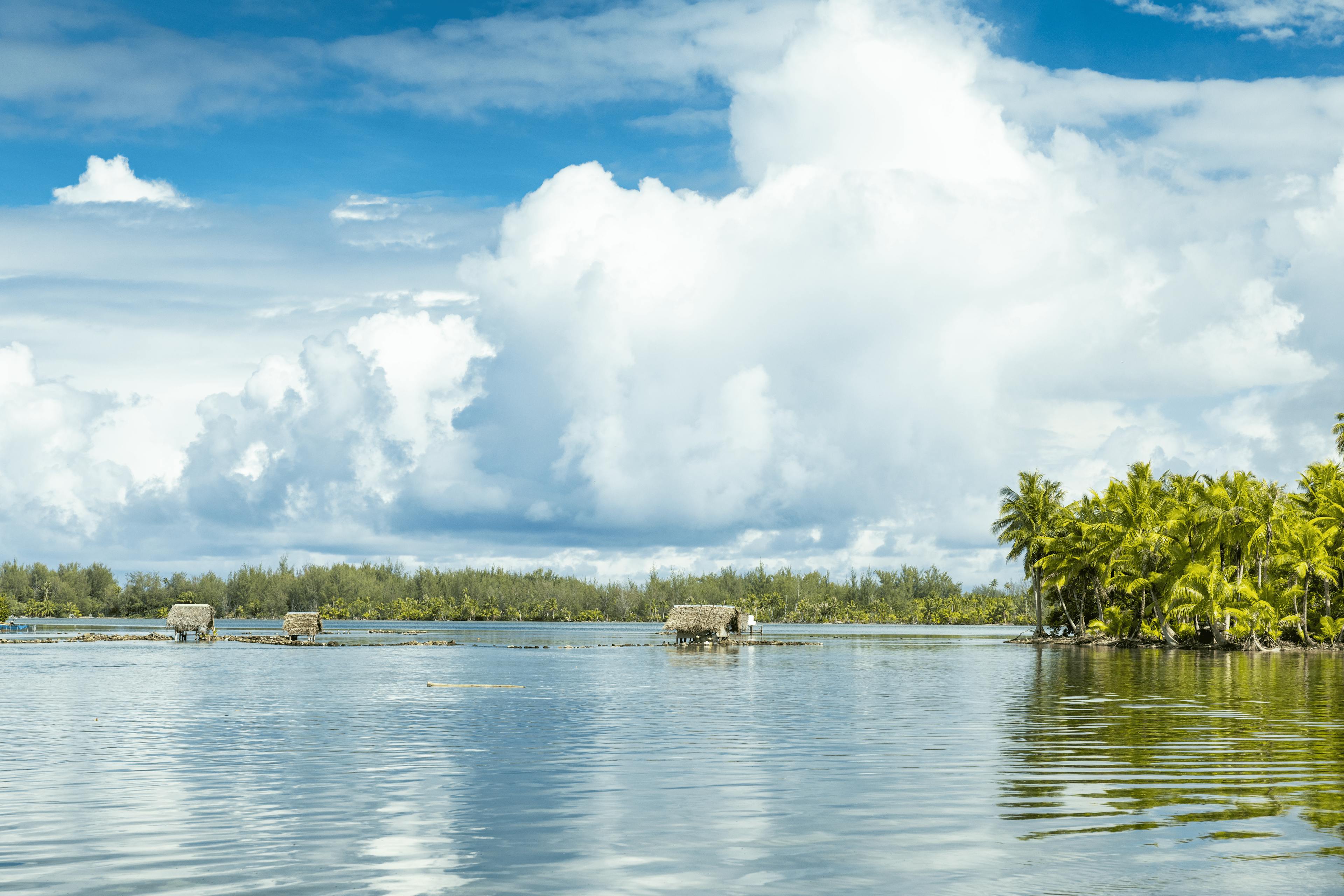 Tranquil scene of Huahine lagoon with two traditional Polynesian fishing huts on calm water, surrounded by lush greenery, under a clear blue sky with fluffy clouds.