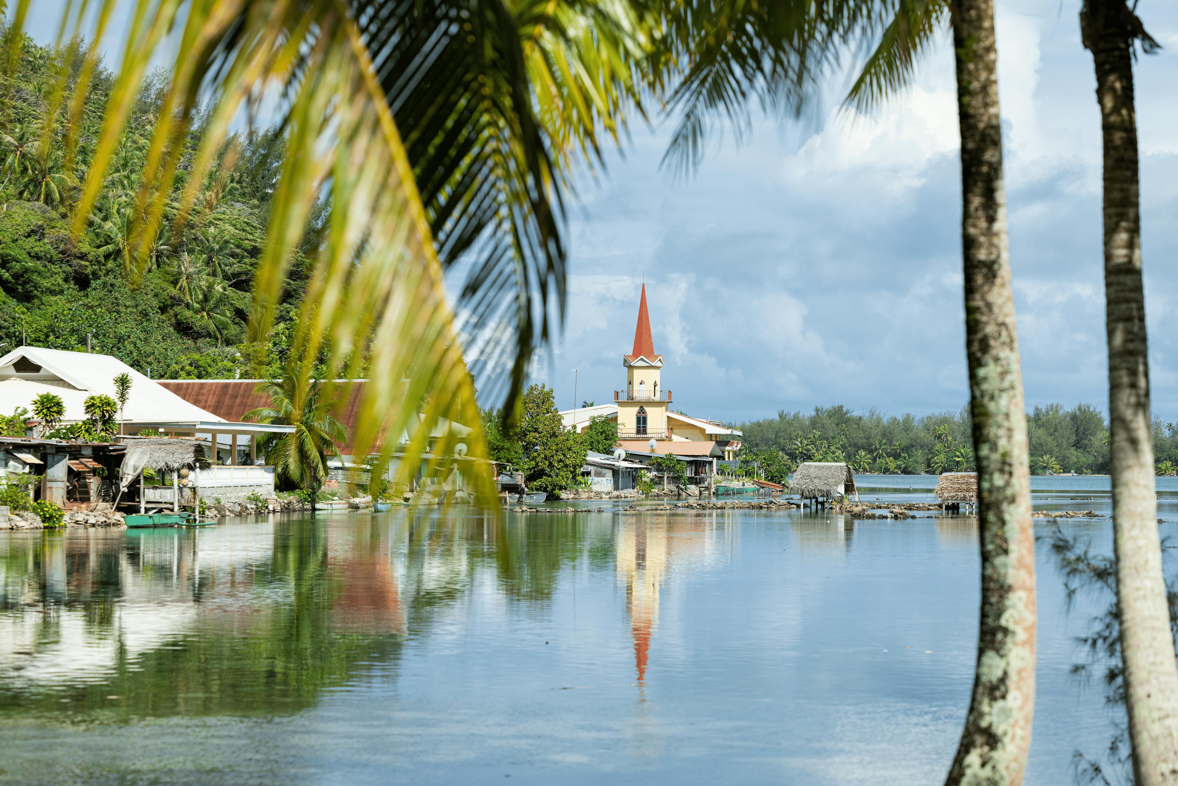 Scenic waterfront scene in Huahine with a distinctive church featuring a bright red spire, reflecting beautifully in the calm lagoon waters, framed by lush tropical foliage.