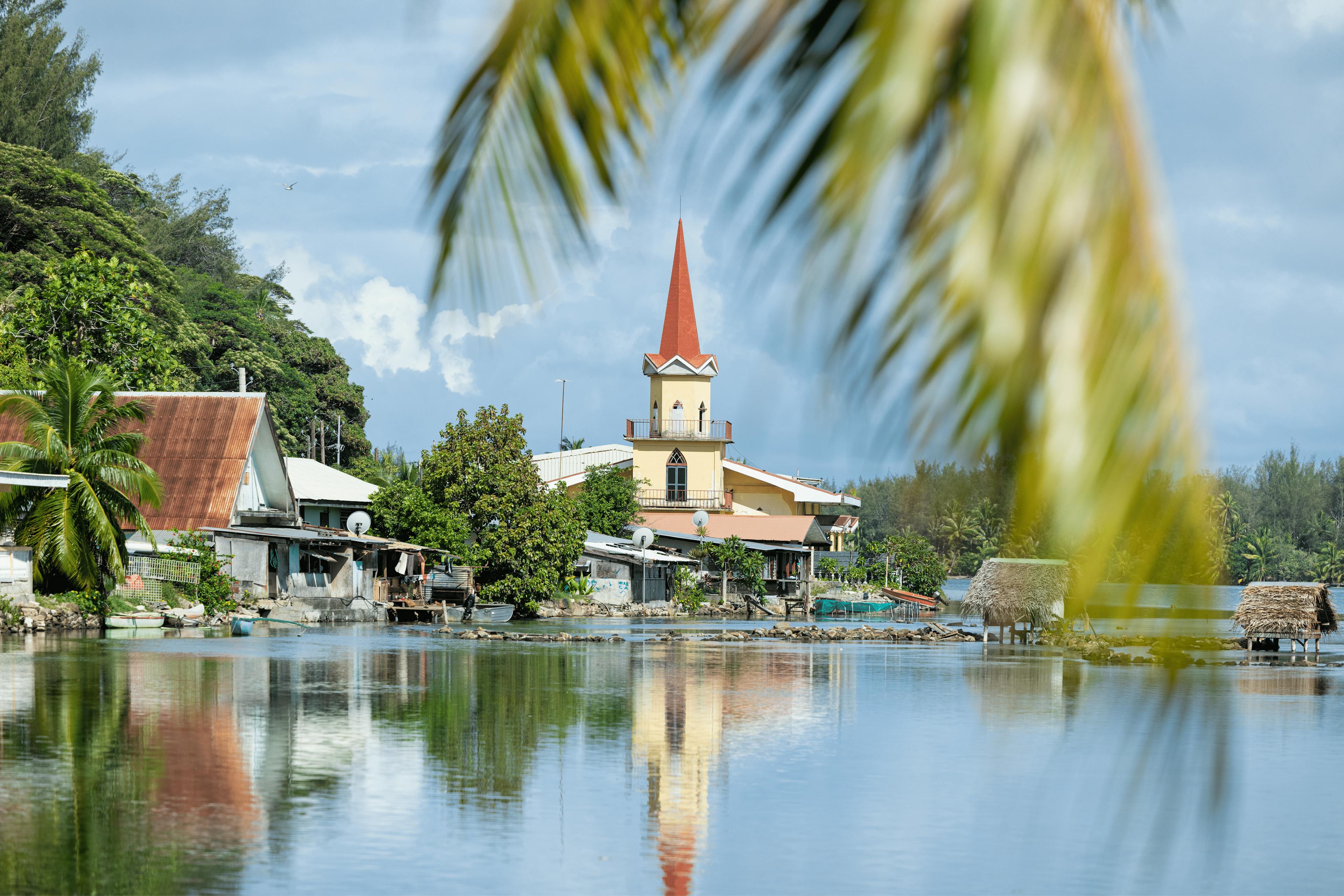 Picturesque view of a small town in Huahine, featuring a charming church with a red roof reflecting in the water, surrounded by tropical palms and clear skies.