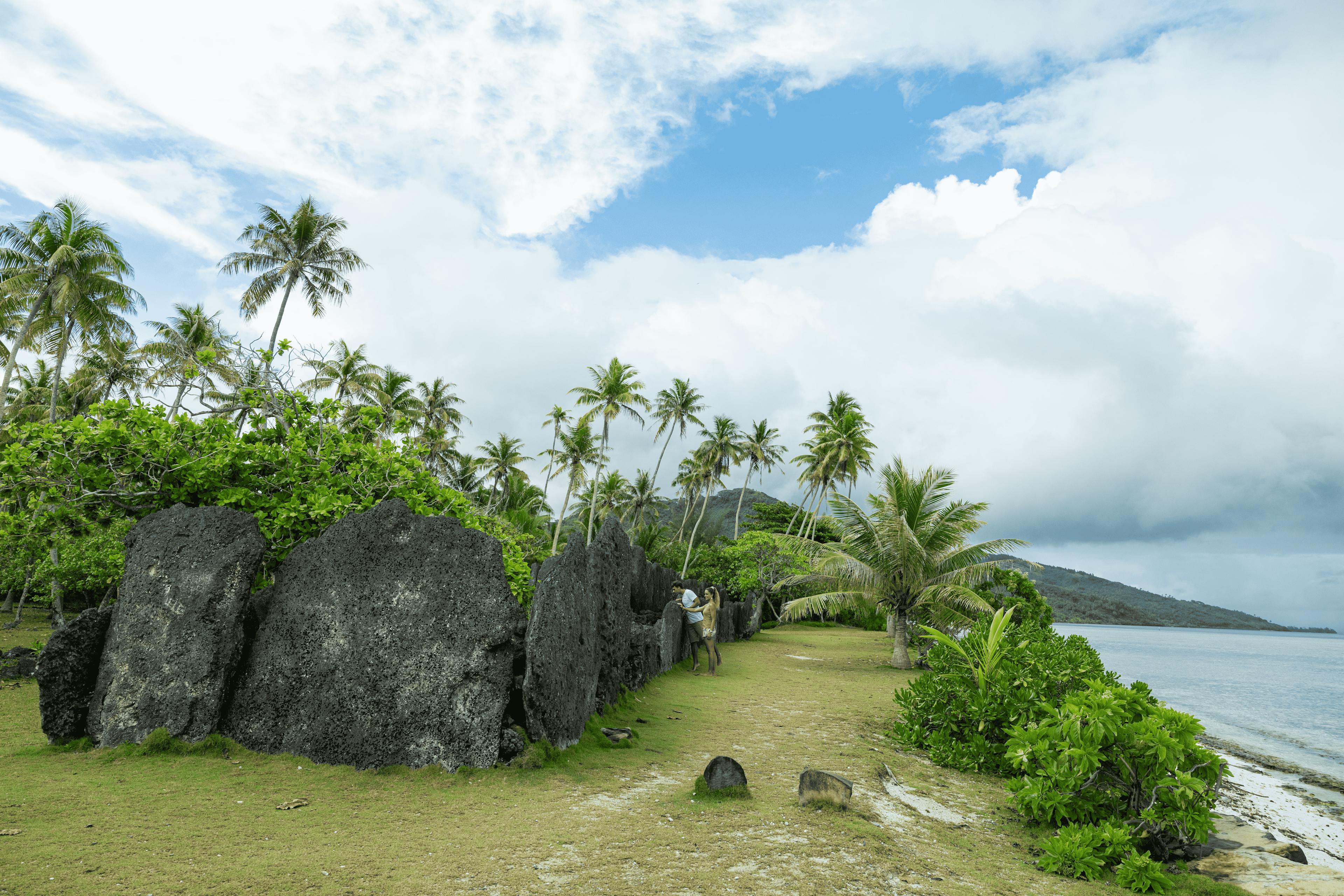 Ancient Polynesian stone formations on a beach in Huahine, surrounded by palm trees and lush foliage, under a cloudy sky.