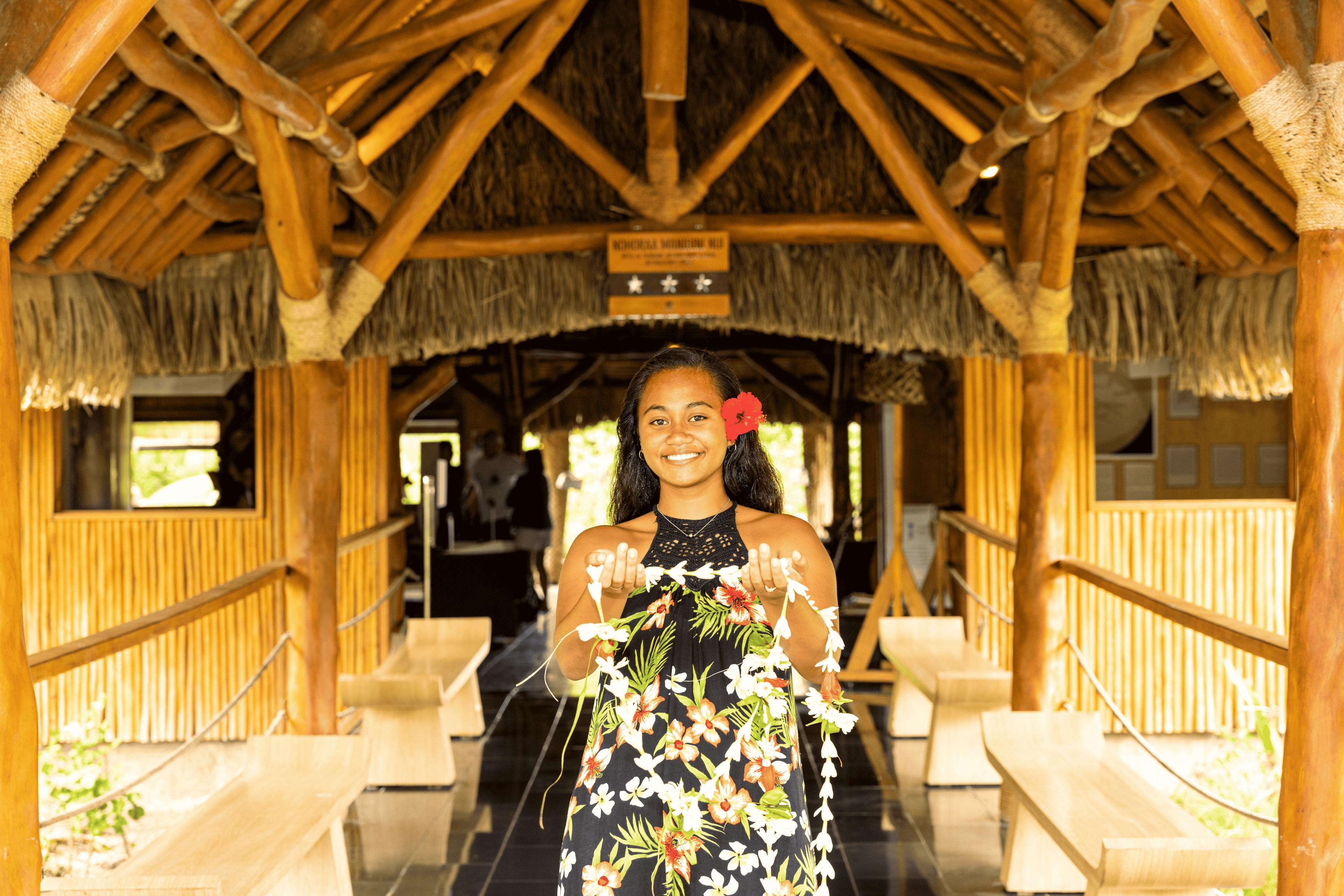 A welcoming Polynesian woman with a bright smile, offering flowers at a traditional bamboo lobby in Huahine, enhancing the local cultural experience.