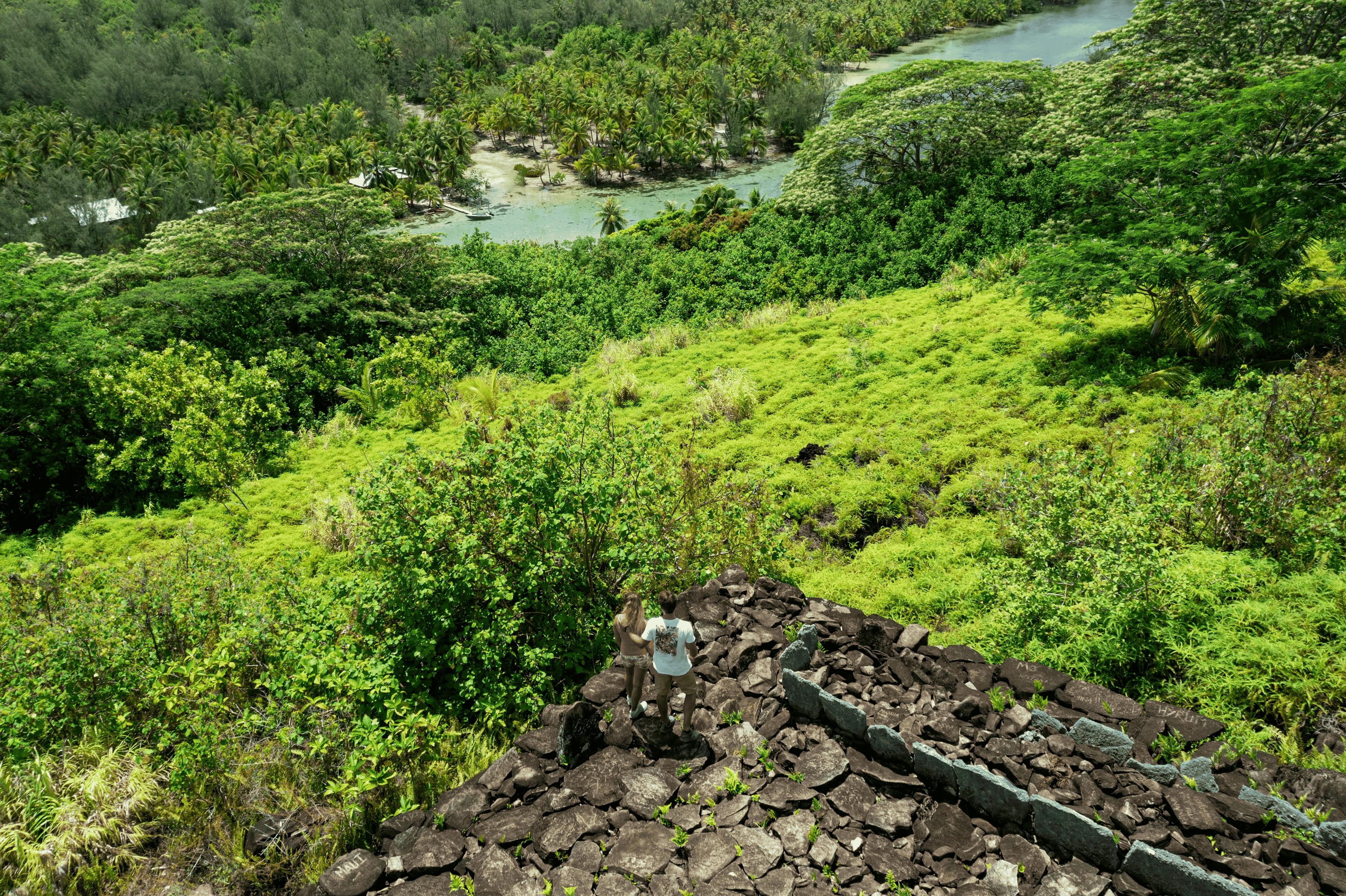 Couple exploring Huahine's natural beauty, standing together on a rocky overlook with a panoramic view of the lush island landscape.