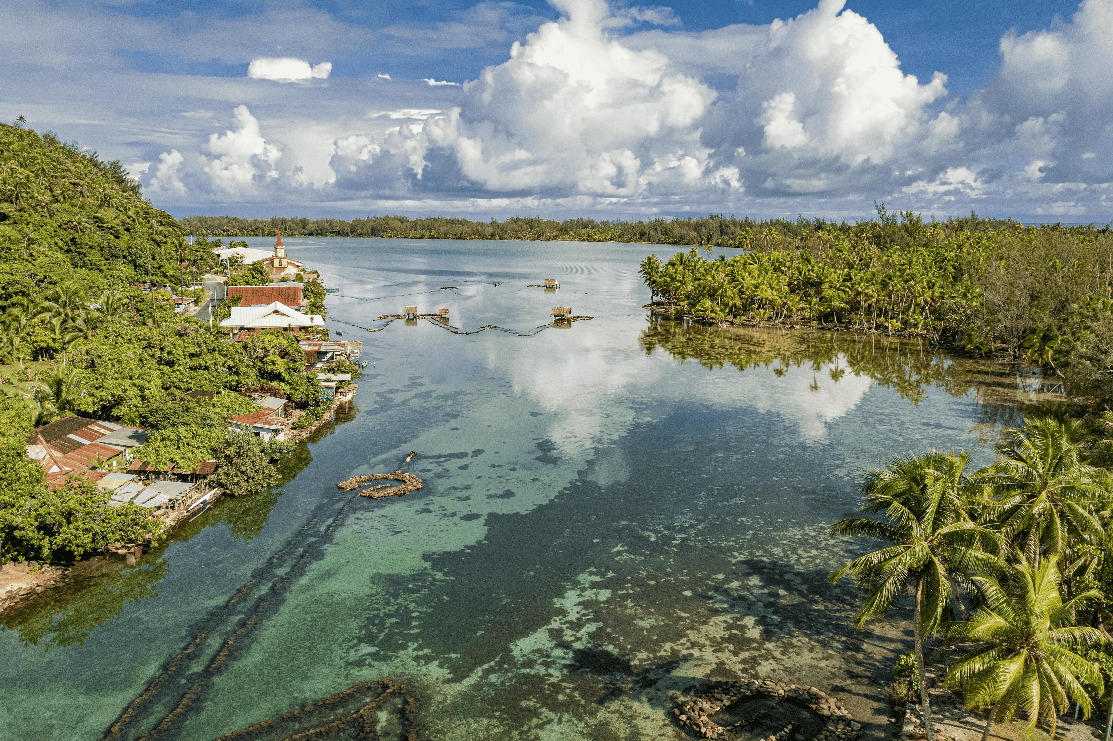 Vue aérienne de Huahine, une île de Polynésie française, montrant un lagon tropical entouré de végétation luxuriante, avec des maisons côtières, des palmiers et des enclos de pêche traditionnels sur l’eau. Le ciel bleu et les nuages blancs se reflètent dans l’eau calme.