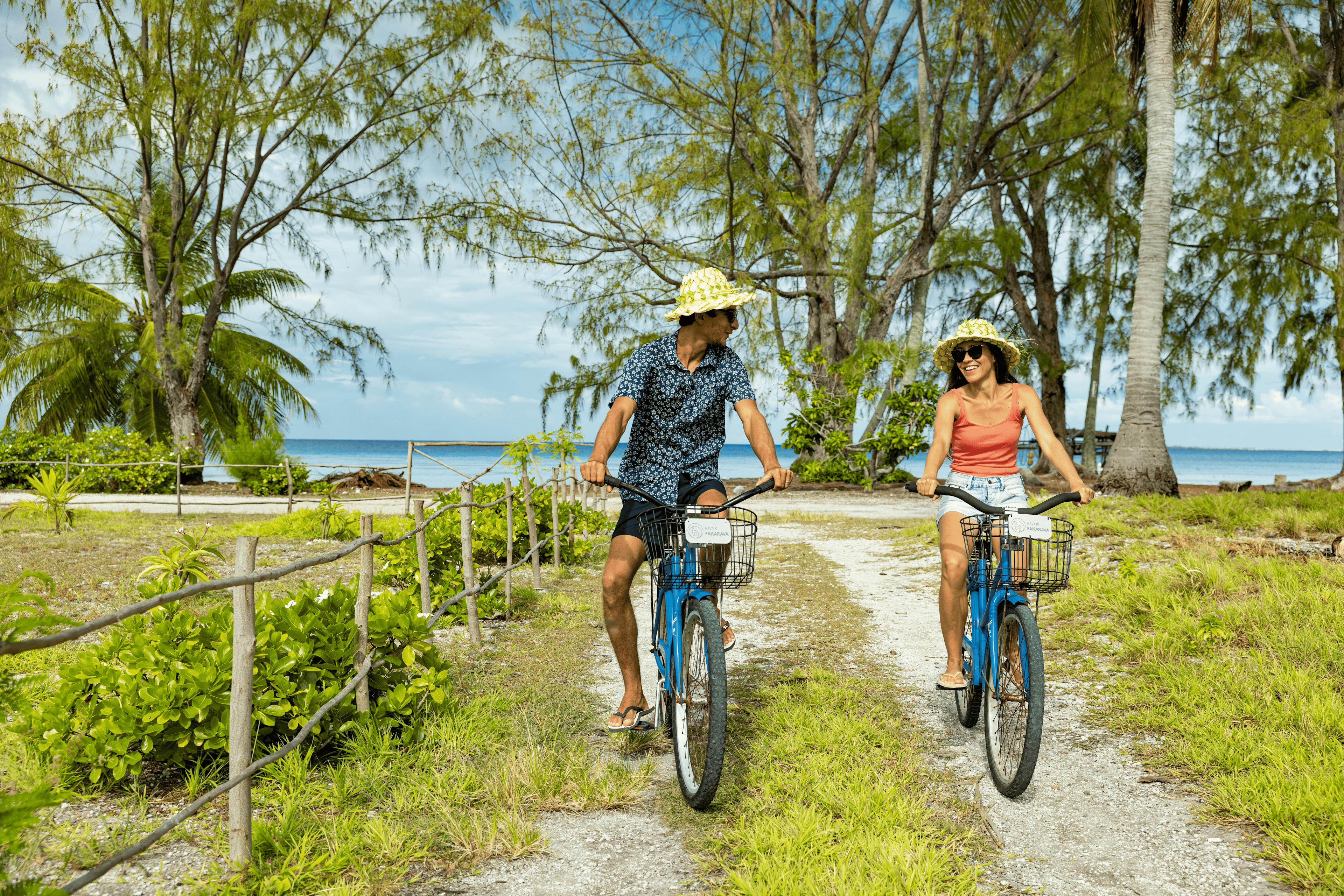 Un couple vêtu de tenues estivales roule à vélo sur un chemin de terre bordé de verdure et de palmiers. Avec la mer turquoise en arrière-plan, ils profitent d’un moment de détente et d’exploration sur l'île paradisiaque de Fakarava.