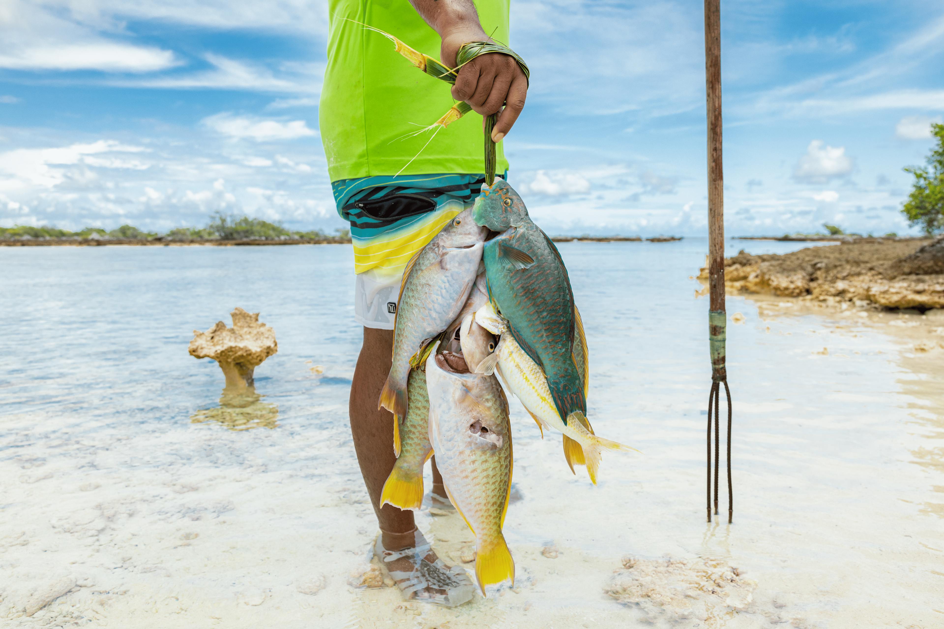 Un pêcheur polynésien tient fièrement une prise de poissons multicolores attachés ensemble avec une fibre végétale. Ses pieds s’enfoncent dans le sable clair alors que l’eau peu profonde du lagon scintille sous le soleil.