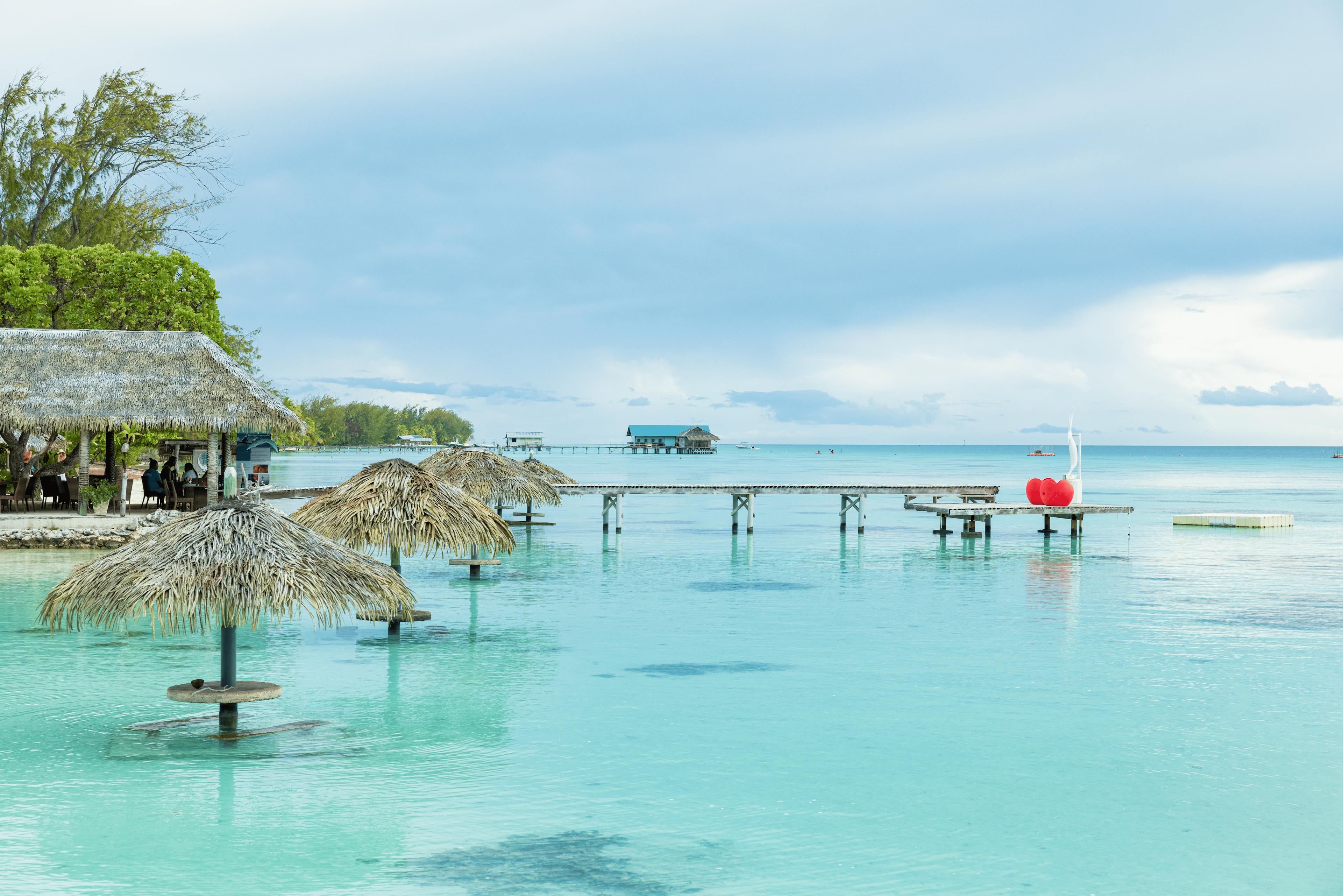 Un restaurant sur pilotis borde le lagon turquoise, avec des parasols en feuilles de palmier émergeant directement de l’eau. Une ambiance paisible règne alors que les visiteurs profitent de la vue sur l’océan et des saveurs locales sous une brise tropicale.