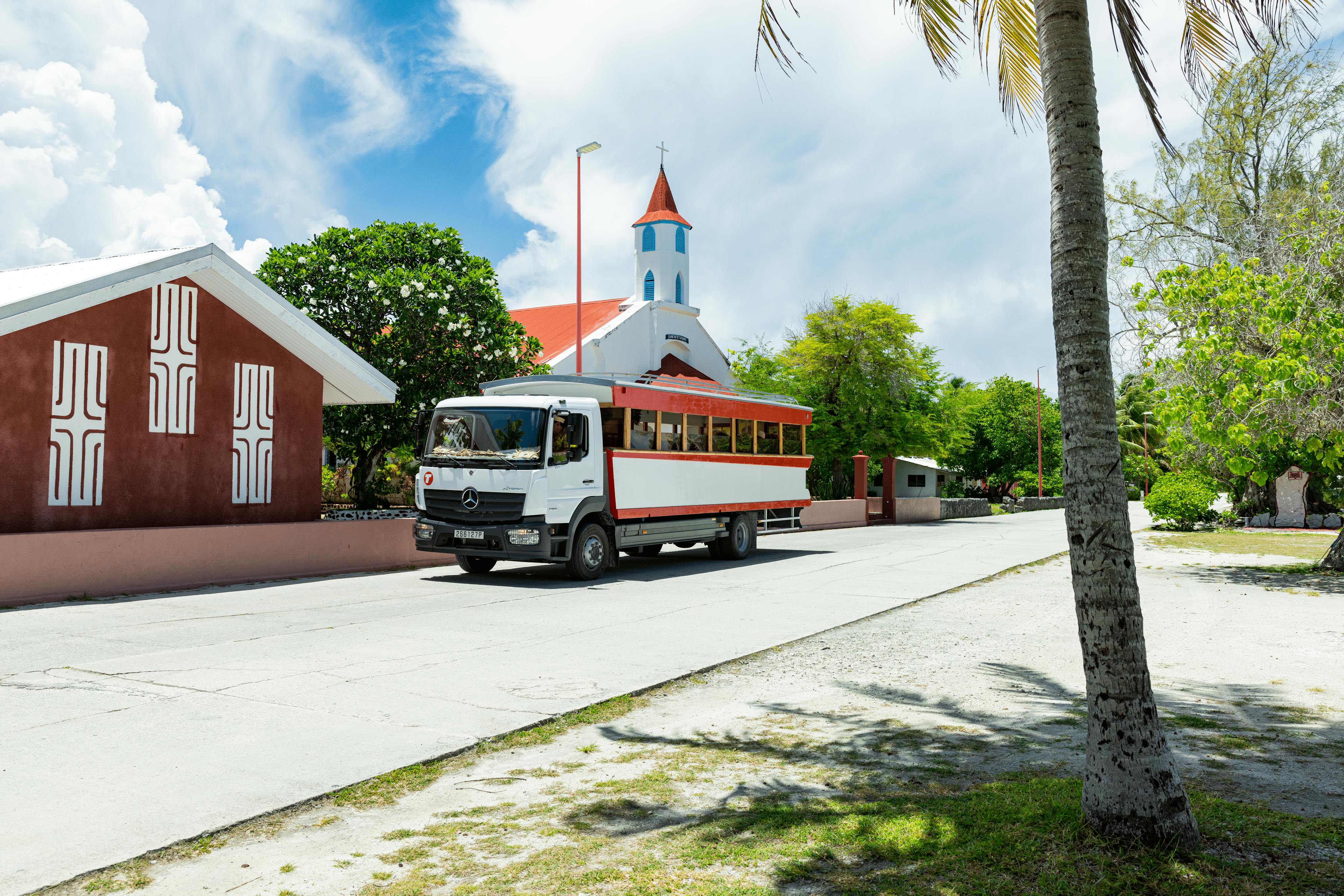 Un bus touristique aux couleurs rouge et blanc circule sur une route bordée de cocotiers à Fakarava. En arrière-plan, une charmante église à toit rouge et murs blancs rappelle l’héritage culturel et religieux de l’île.