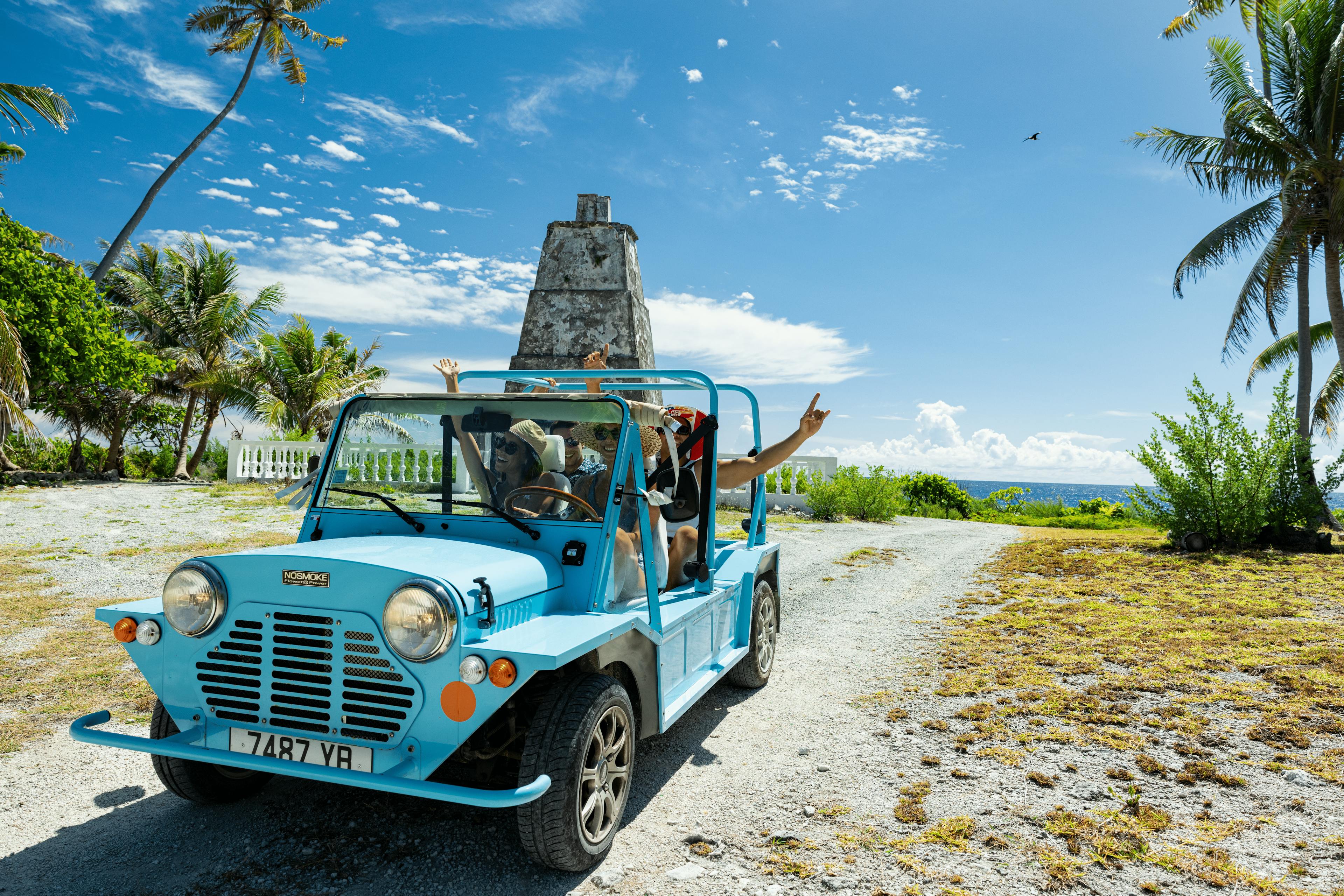 Une voiture de plage décapotable bleu ciel roule sur un chemin bordé de cocotiers à Fakarava. Les passagers, coiffés de chapeaux de paille, lèvent les bras avec enthousiasme alors qu’ils s’approchent d’un ancien monument en pierre face à l’océan.