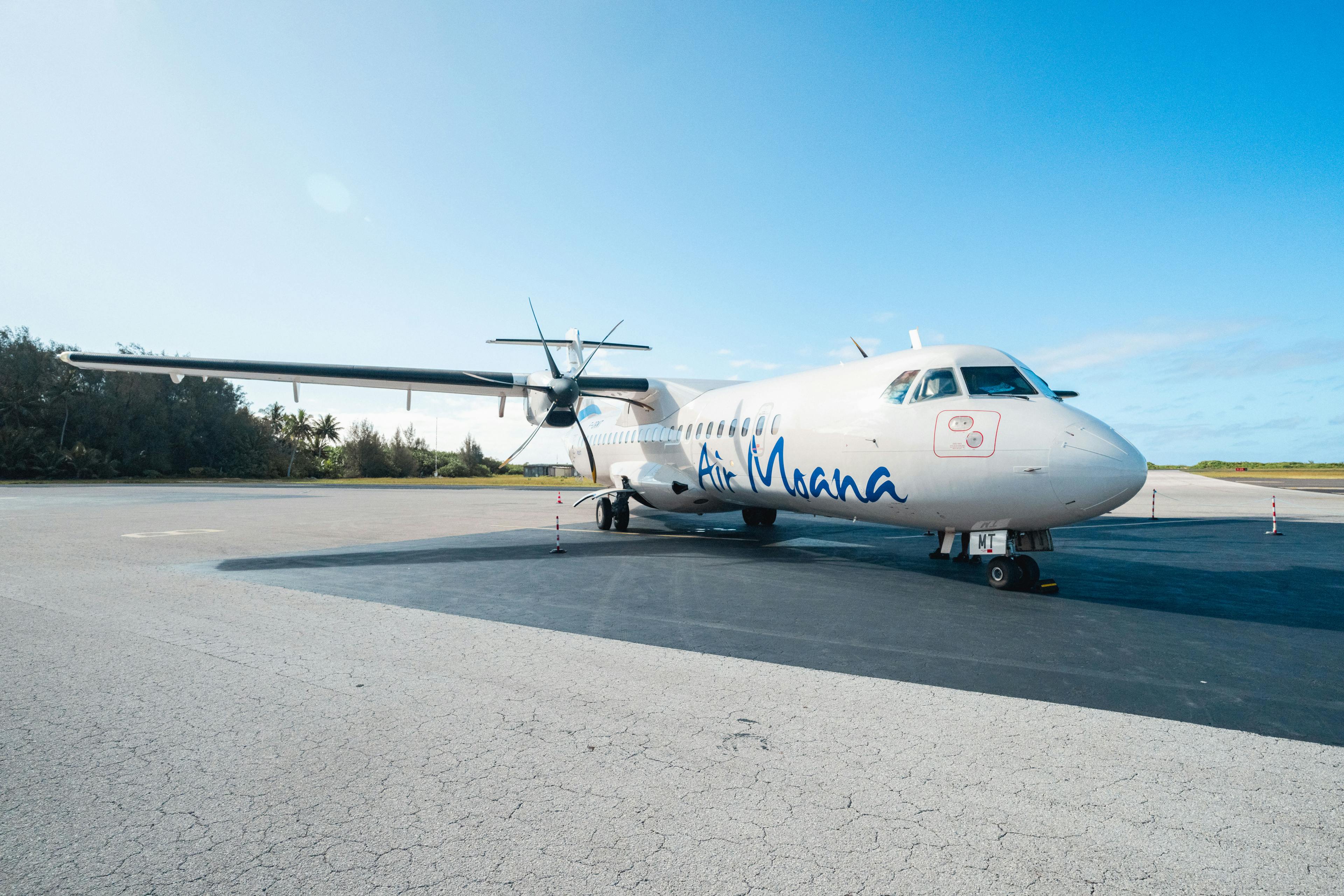 Un avion Air Moana vue de l'avant sur le tarmac avec un ciel bleu et des sapins en arrière plan