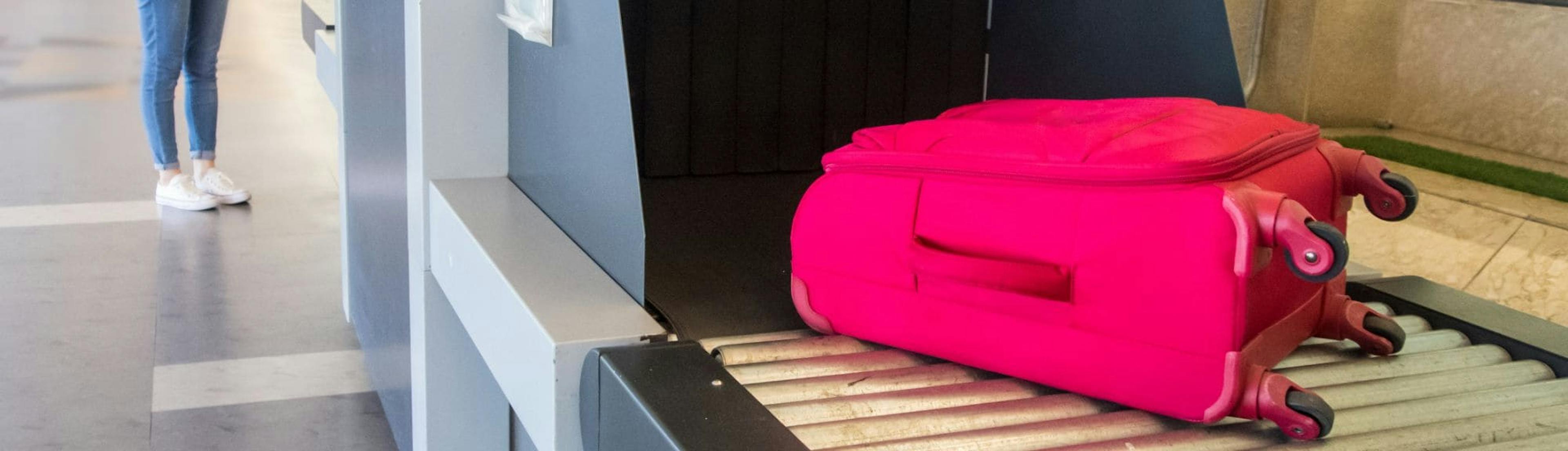 A Chinese woman waits next to an X-ray baggage scanner at the airport, watching her bright pink suitcase go through on the conveyor belt. She is wearing a plaid shirt, jeans, and a backpack.