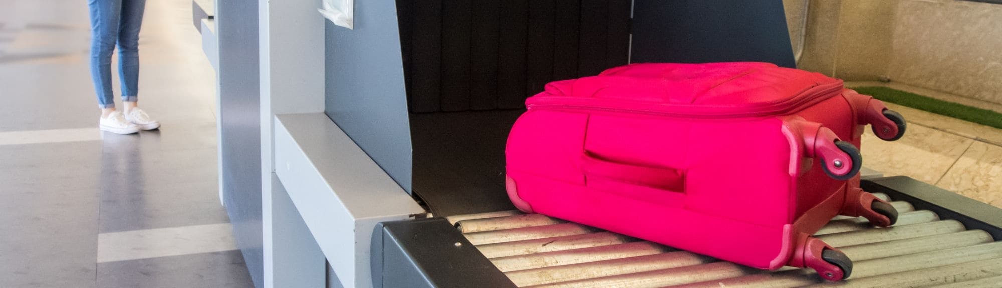 Une femme chinoise attend à côté d'un scanner à rayons X pour les bagages à l'aéroport, regardant sa valise rose vif passer sur le tapis roulant du scanner. Elle porte une chemise à carreaux, un jean, et un sac à dos.