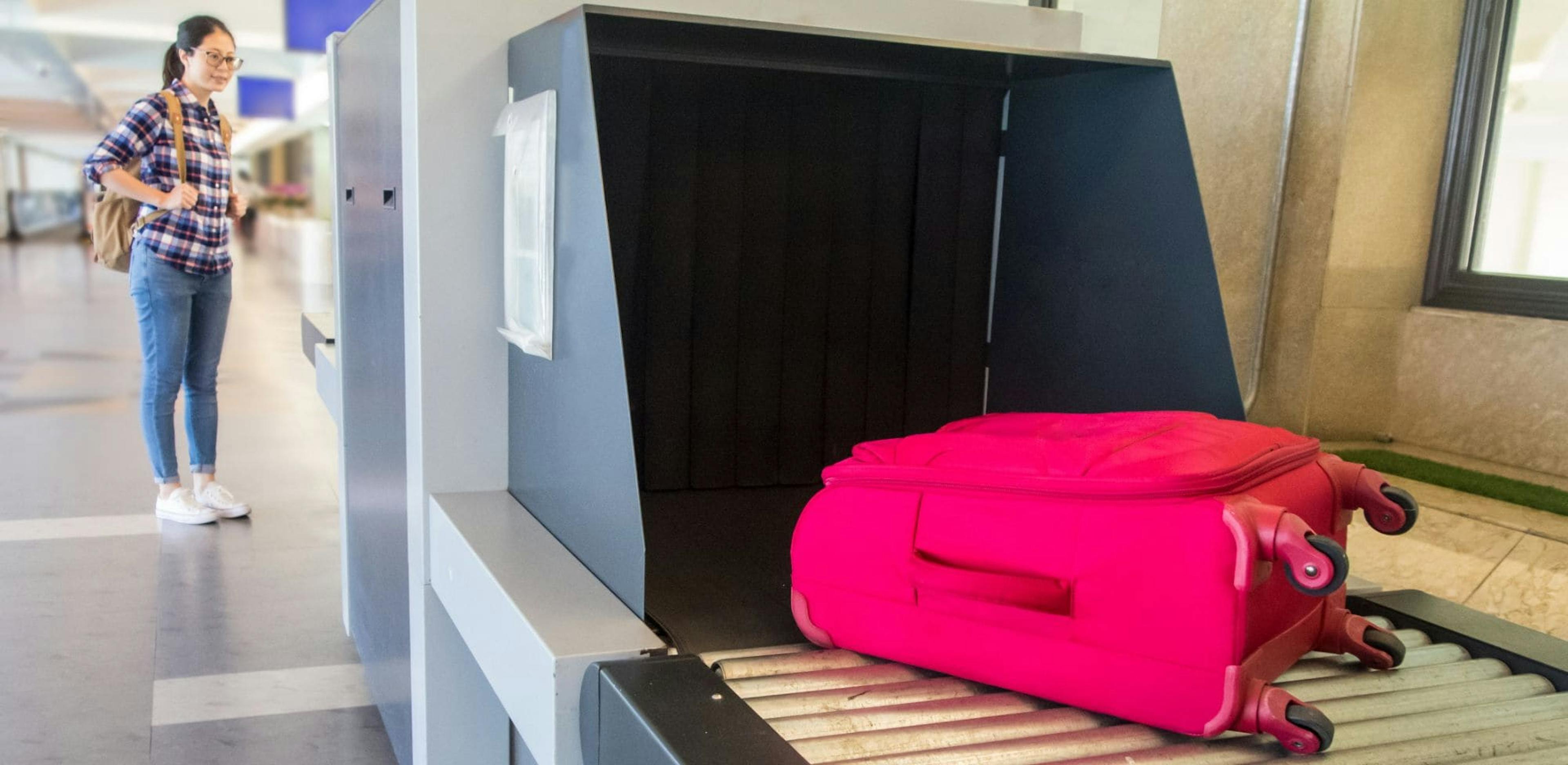 A Chinese woman waits next to an X-ray baggage scanner at the airport, watching her bright pink suitcase go through on the conveyor belt. She is wearing a plaid shirt, jeans, and a backpack.