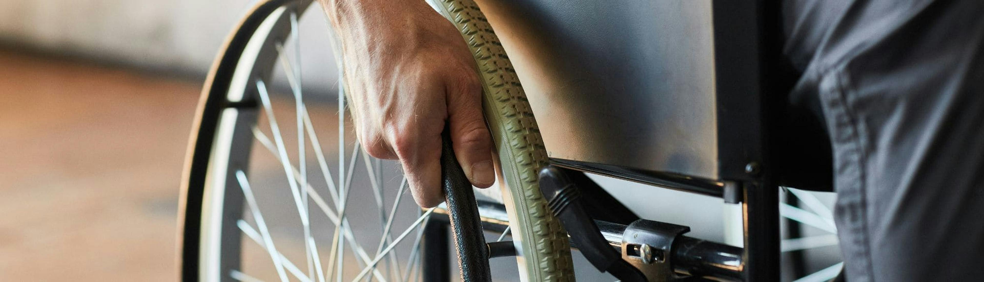 Close-up view of a man in a wheelchair, holding the wheel with his hand to move. The image focuses on the hand and side of the wheelchair, emphasizing independence and mobility.