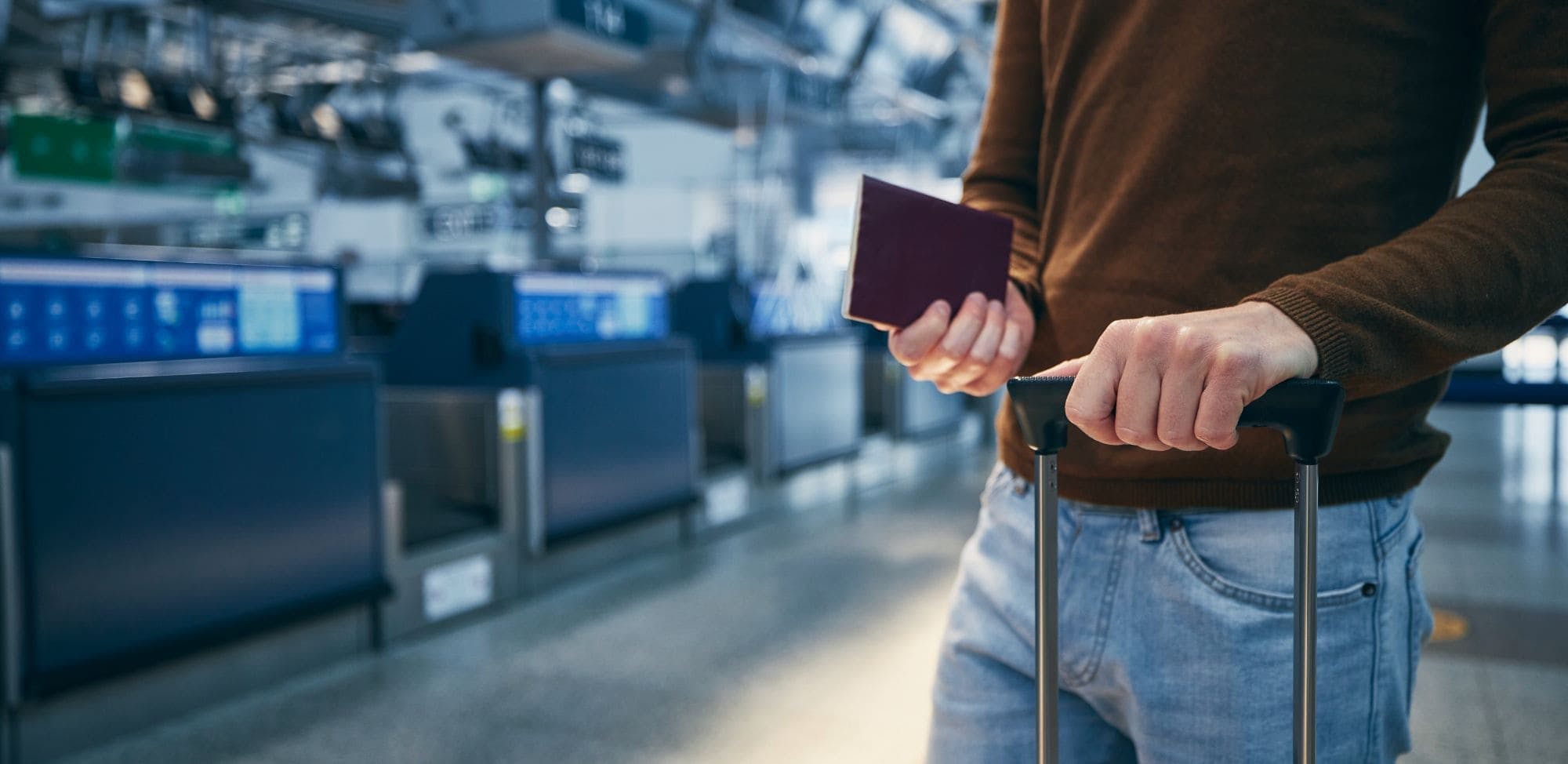 Un passager se tient devant un comptoir d'enregistrement à l'aéroport, tenant une valise et un passeport dans ses mains. L'homme porte un pull marron et un jean. Le fond montre un espace aéroportuaire flou avec des écrans et des comptoirs d'enregistrement.